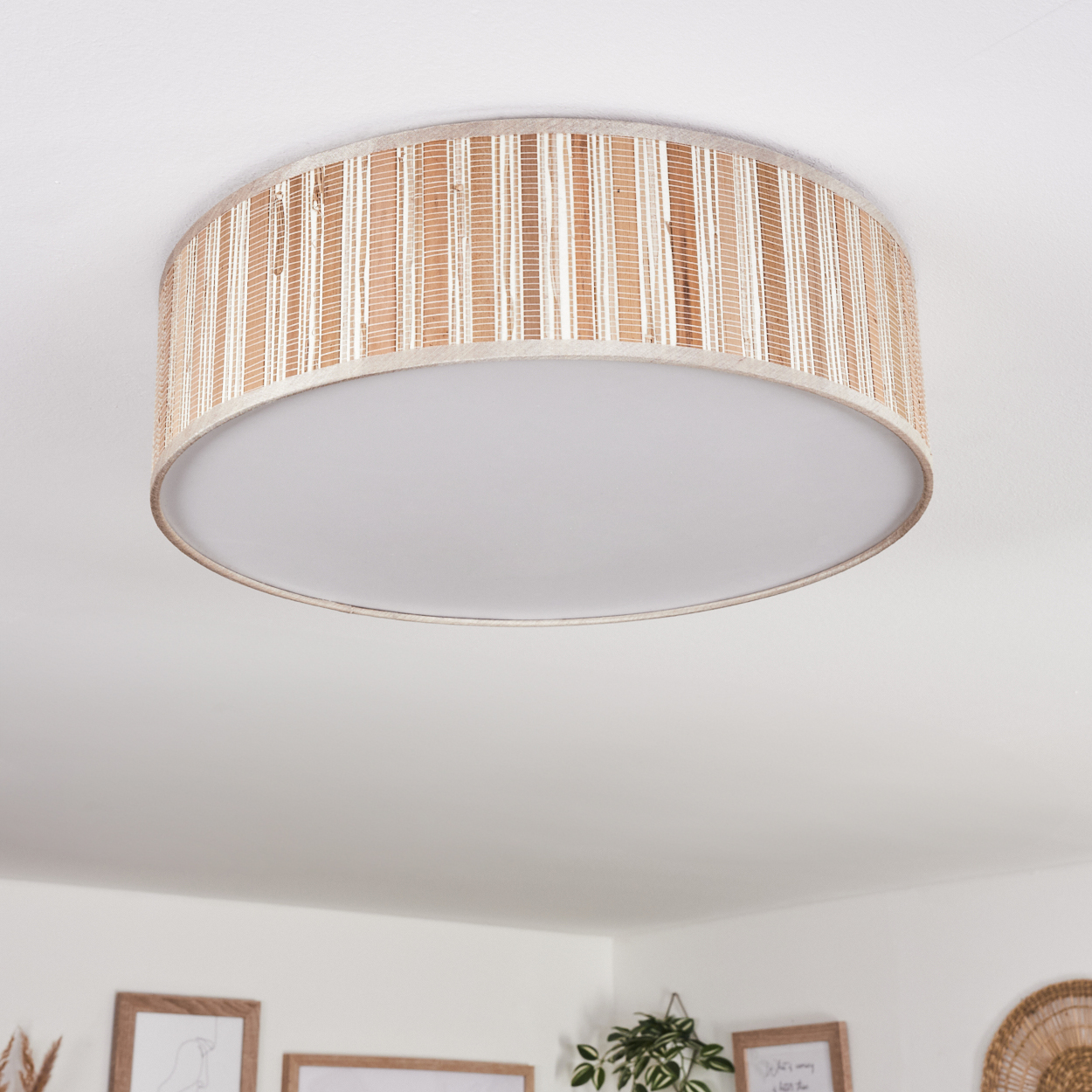 Eine runde Deckenlampe mit gewebtem, beige-weißem Schirm hängt an einer weißen Decke. Kunstwerke und Pflanzen sind unten sichtbar.