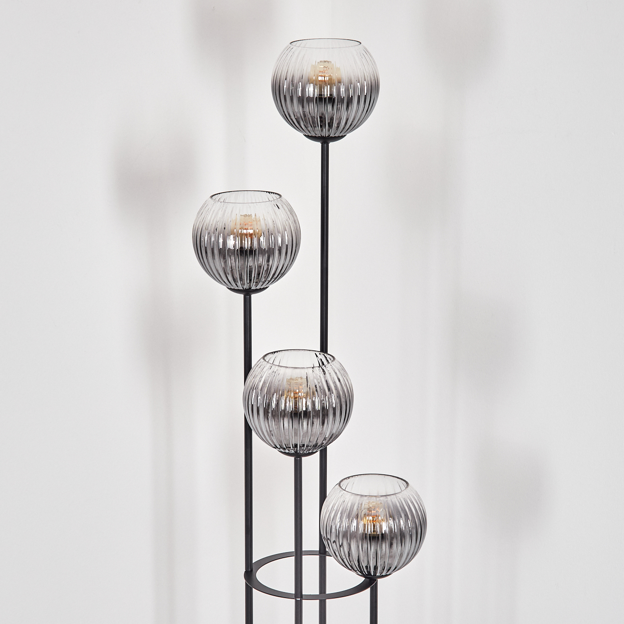 Eine moderne Stehlampe mit vier Glasschirmen, jeweils mit einer Glühbirne. Der Metallrahmen ist schwarz, der Hintergrund eine weiße Wand.
