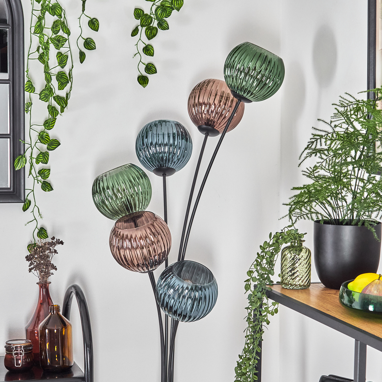 Eine dekorative Stehlampe mit bunten Glaskugeln. Rechts grüne Pflanzen und ein schwarzer Topf. Zusätzliche Artikel auf einem Regal.