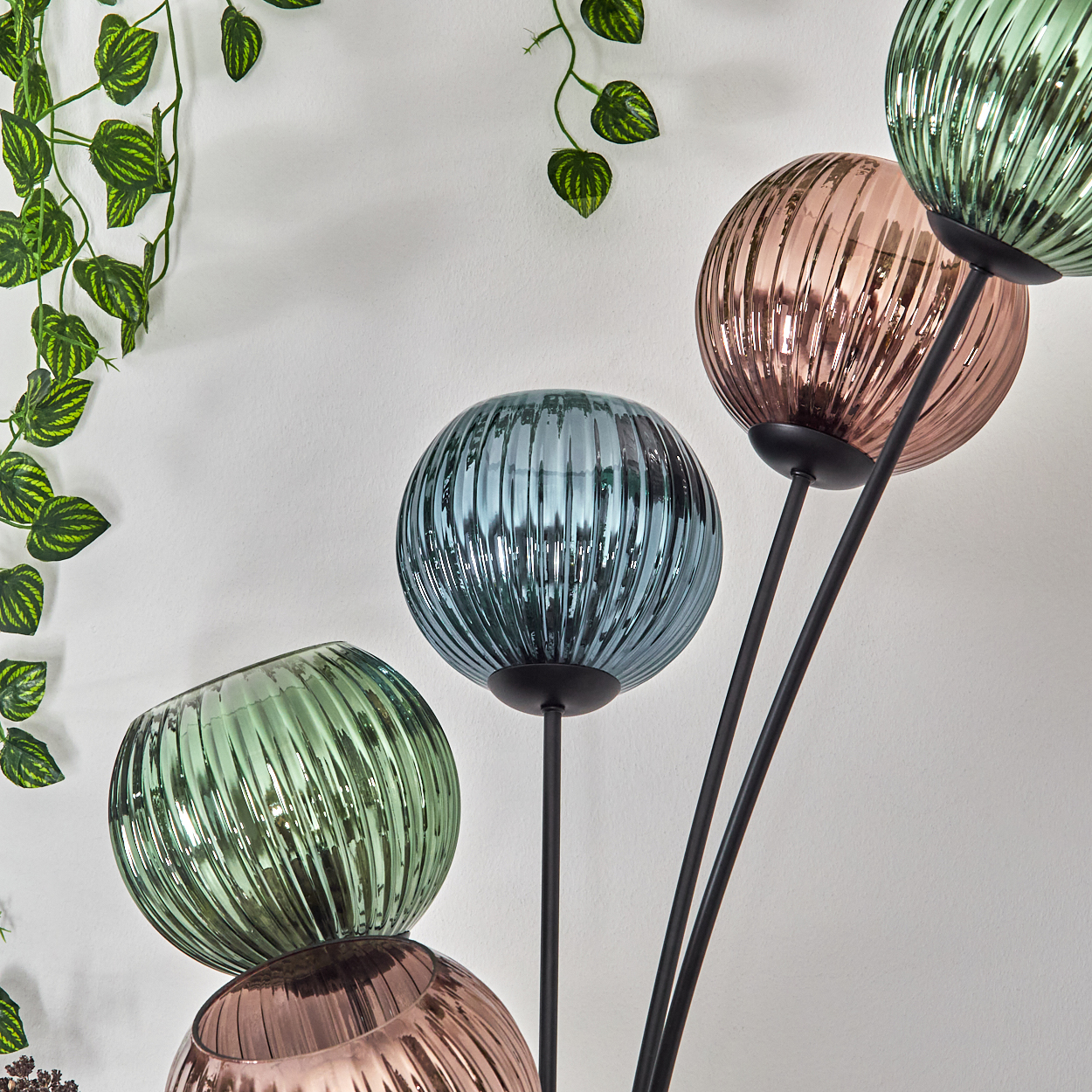 Bunte Glaskugelleuchten auf schwarzen Ständern. Grüne, blaue und rosa Kugeln vor einer weißen Wand mit hängenden grünen Blättern.
