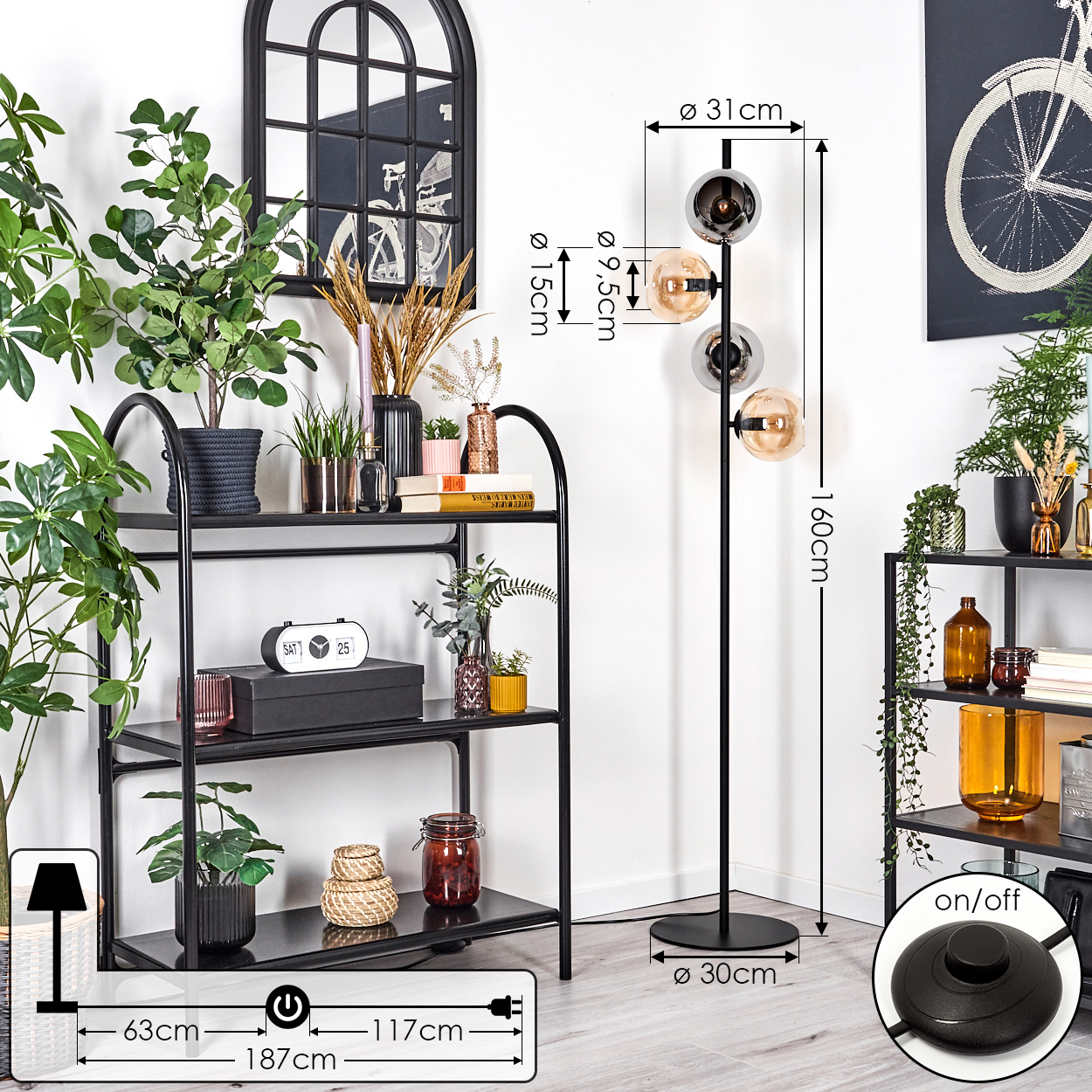Ein Raum mit einer schwarzen Stehlampe mit drei Glaskugeln, einem schwarzen Regal, Pflanzen und einem schwarz gerahmten Spiegel.