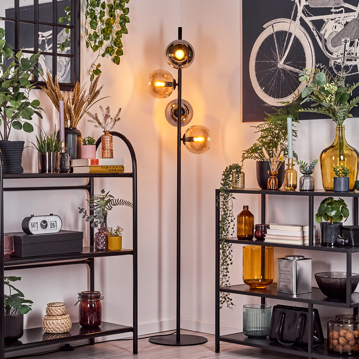 Ein Raum mit Regalen voller Pflanzen und Dekoration. In der Mitte steht eine Stehlampe mit kugelförmigen Lichtern. Ein Fahrradkunstwerk hängt.