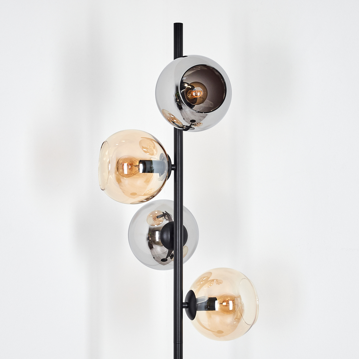 Eine moderne Stehlampe mit vier kugelförmigen Leuchten. Die Schirme sind bernsteinfarben und chromfarben, mit sichtbaren Glühbirnen. Schwarze Stange vor weißer Wand.