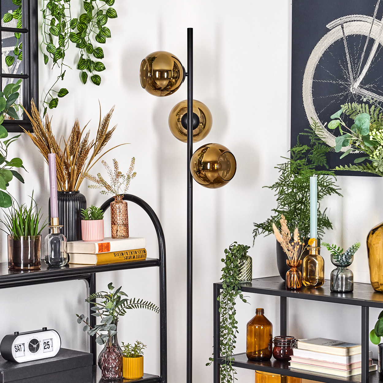 Ein Raum mit einem schwarzen Metallregal, das mit Pflanzen und Vasen dekoriert ist. Eine Stehlampe mit drei goldenen Glühbirnen steht hoch.