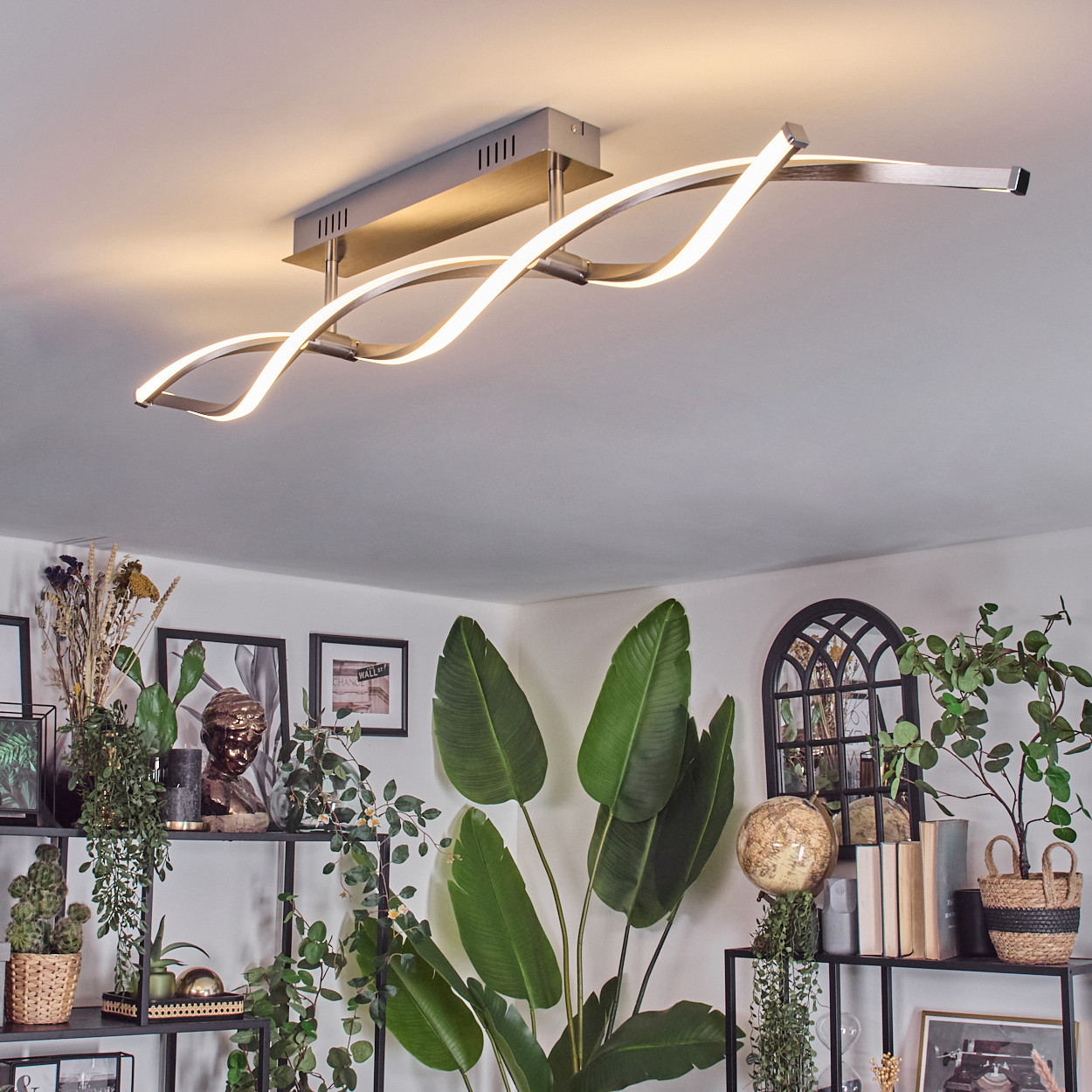Eine moderne Deckenleuchte mit wellenförmigem Design ist eingeschaltet. Darunter dekorieren Pflanzen, Bilder und ein Globus den Raum.