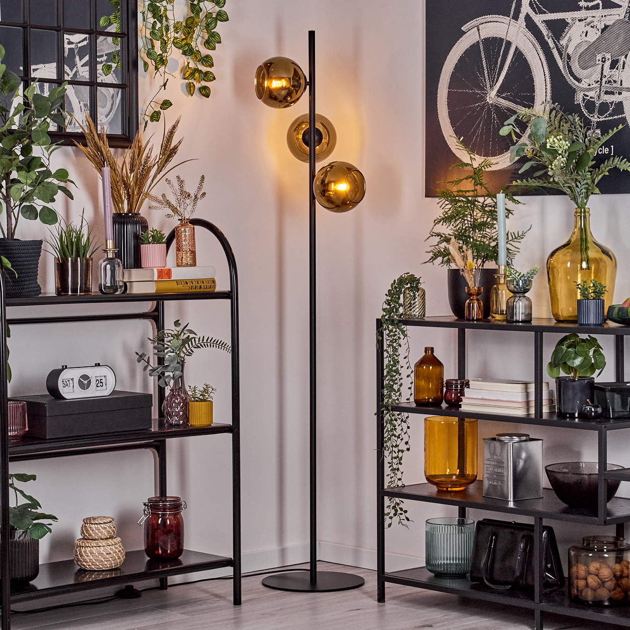 Wohnzimmer mit Regalen, Pflanzen und einer Stehlampe mit drei kugelförmigen Lichtern, Fahrradkunst an der Wand.