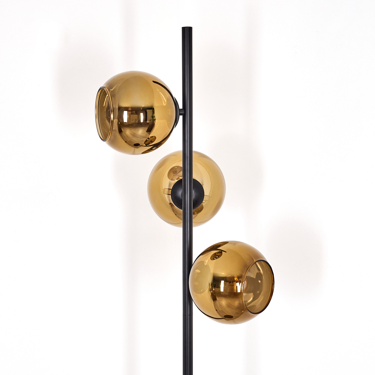 Eine moderne Stehlampe mit drei goldenen Glaskugelschirmen an einer schwarzen Stange, vor einer weißen Wand.