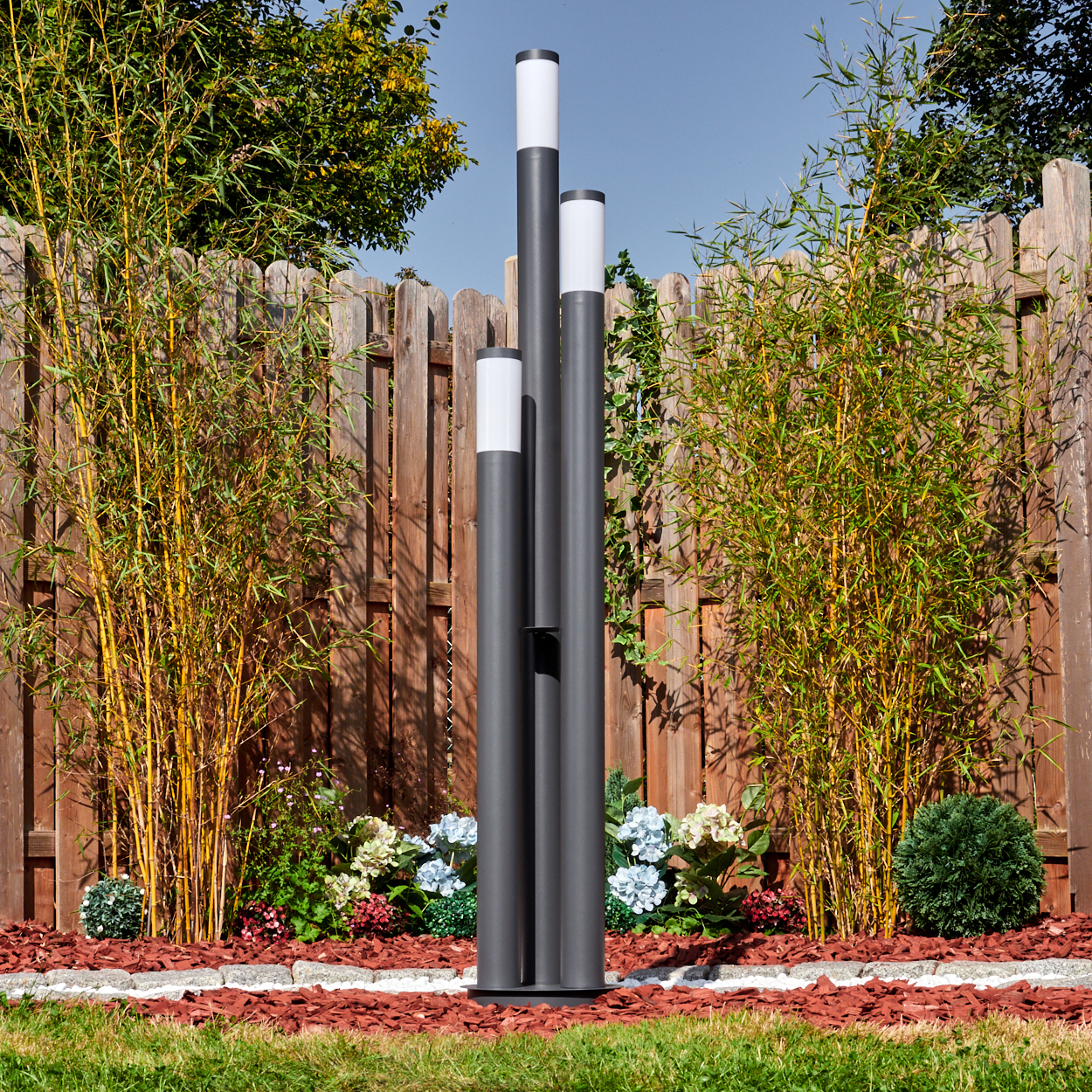 Drei dunkelgraue Außenlampen stehen in einem Garten mit Holzzaun, Bambus und Blumen. Der Himmel ist blau.