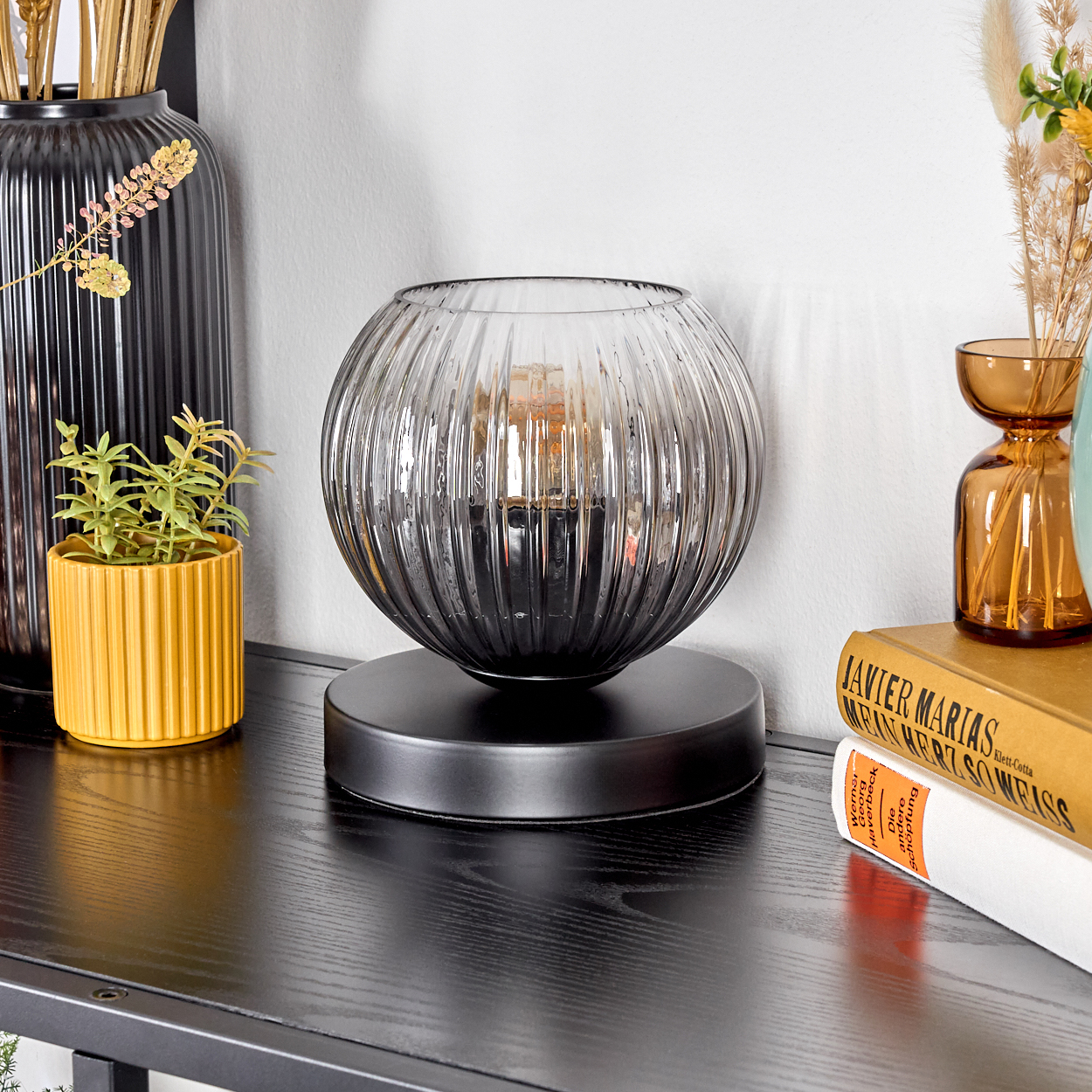 Eine dekorative Lampe mit geripptem Glasschirm steht auf einem schwarzen Tisch. Weitere Dekorationen sind Vasen, Bücher und Topfpflanzen.