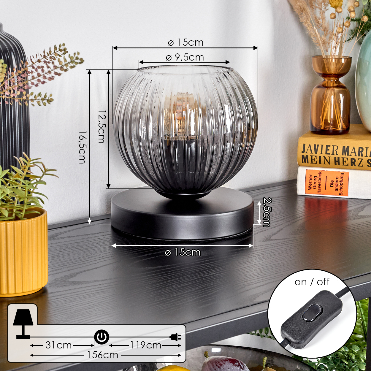 Eine dekorative Tischlampe mit dunklem Glasschirm steht auf einem schwarzen Tisch. Bücher und Vasen sind im Hintergrund. Ein Ein-/Ausschalter ist sichtbar.
