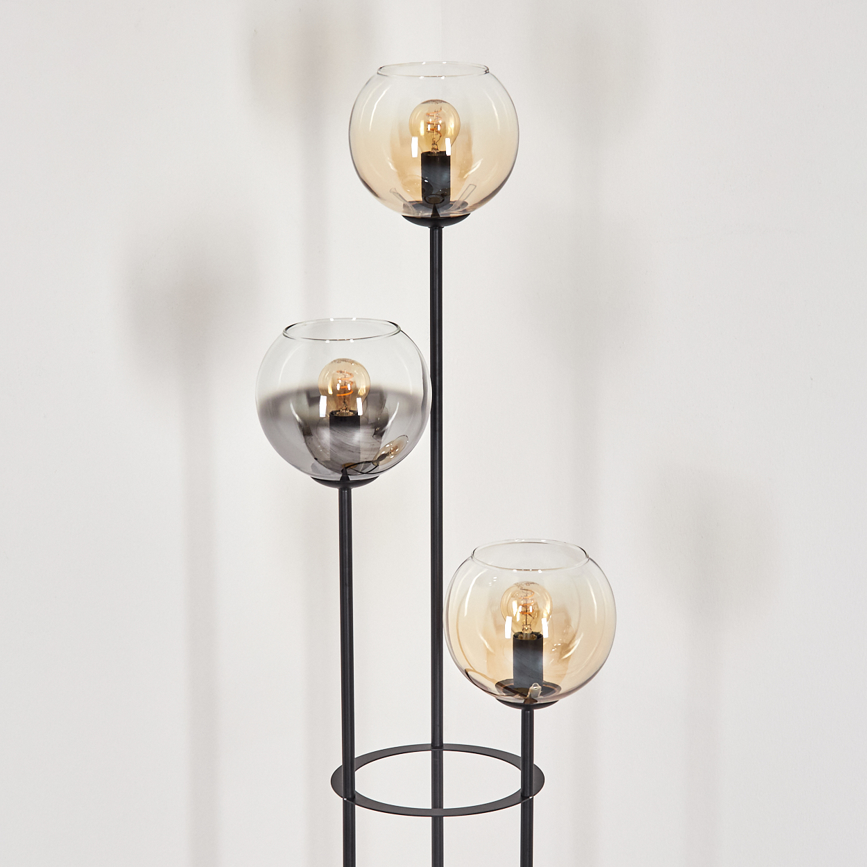 Eine moderne Stehlampe mit drei kugelförmigen Lampenschirmen. Jeder Schirm hat eine Glühbirne. Schwarzer Metallrahmen. Hintergrund ist eine weiße Wand.