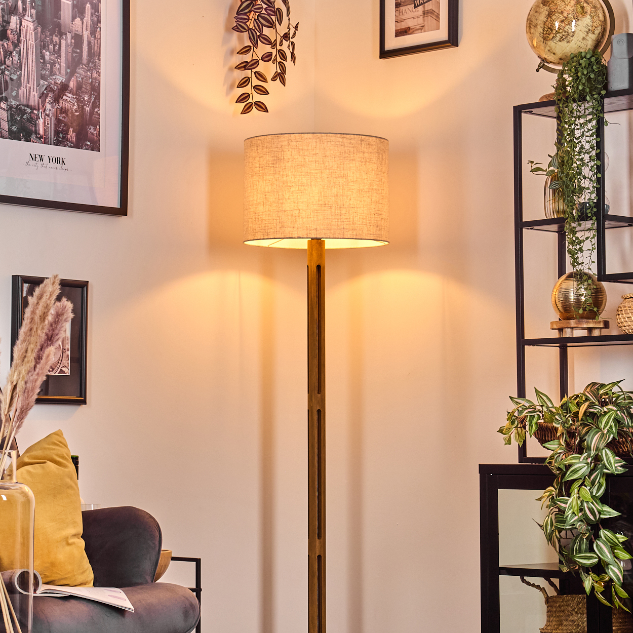 Eine beleuchtete Stehlampe steht in einem Raum mit einem Regal voller Pflanzen, einem gerahmten New York-Foto und einem Sofa mit gelbem Kissen.