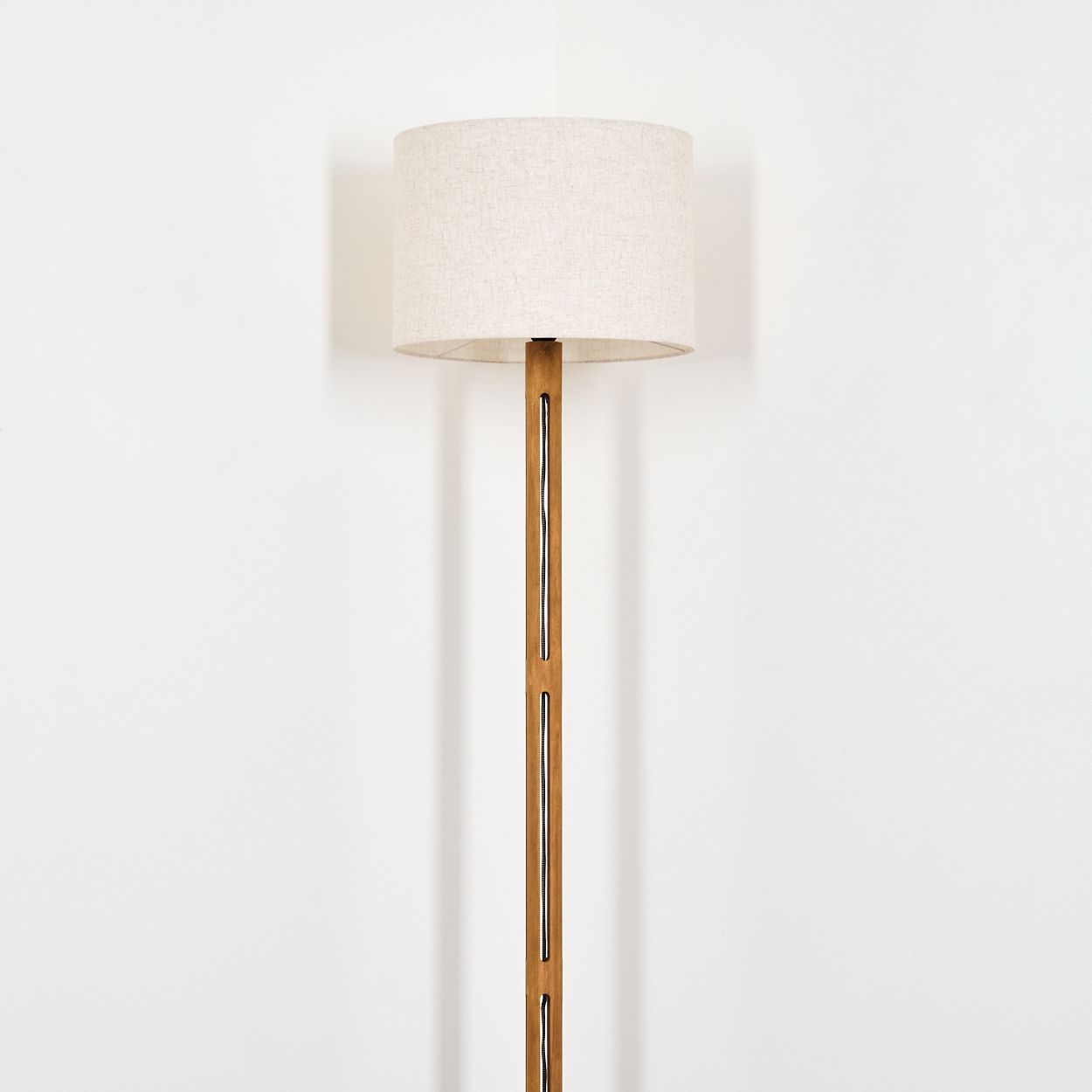 Ein beiger Lampenschirm sitzt auf einer Holzlampe. Die Lampe hat ein rechteckiges Design mit einer Schnur in der Mitte. Weißer Wandhintergrund.