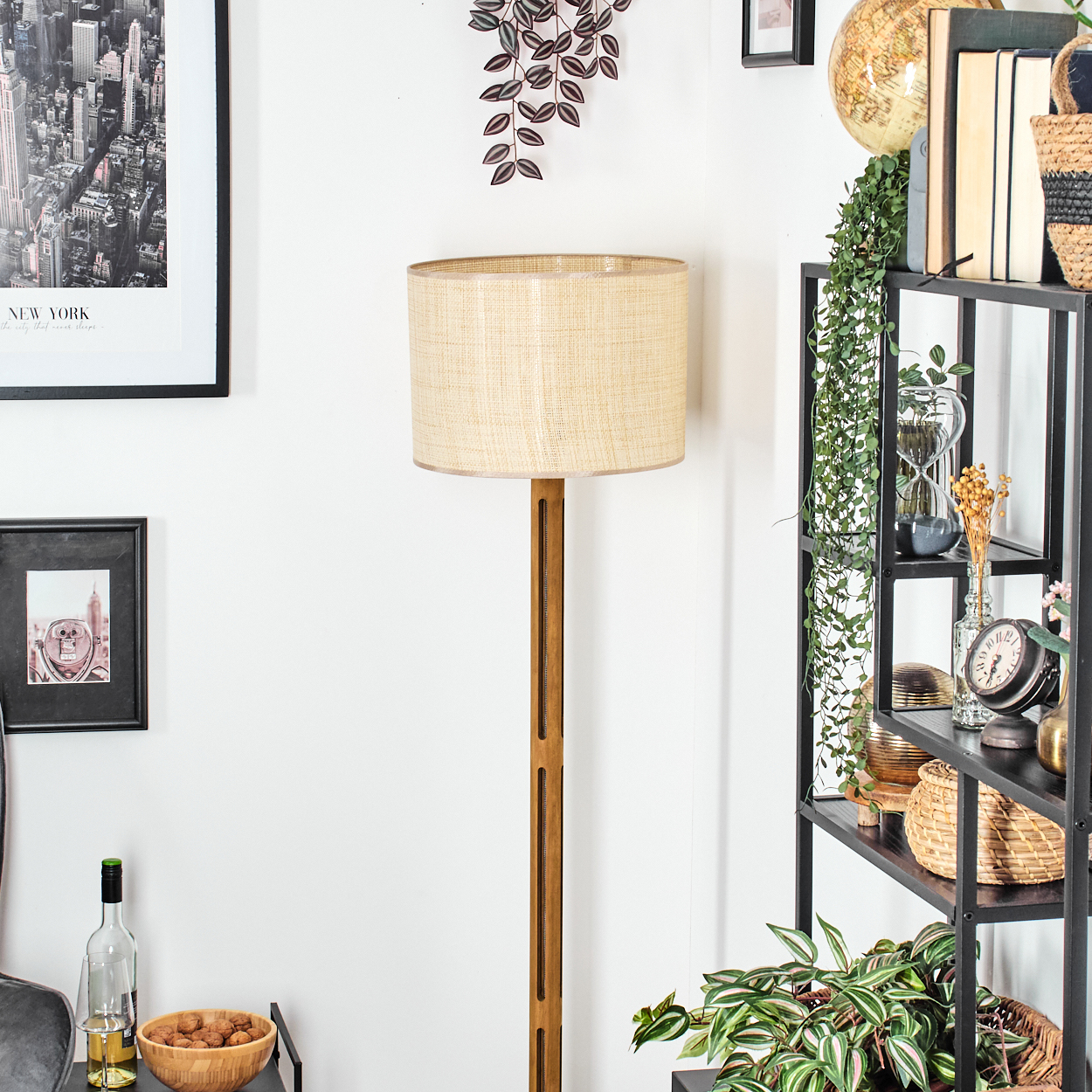 Ein Raum mit Lampe, Bücherregal, gerahmten Bildern und Pflanzen. Die Lampe hat einen beigefarbenen Schirm. Das Regal enthält Bücher, einen Globus und Dekoration.