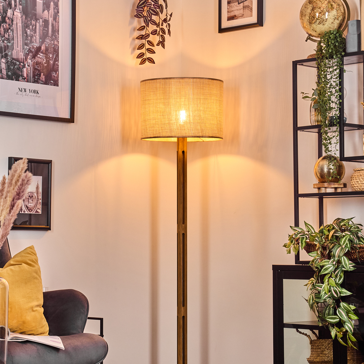 Eine beleuchtete Stehlampe steht in einem Raum mit gerahmten Bildern und einem Bücherregal. Ein gelbes Kissen liegt auf einem Stuhl.