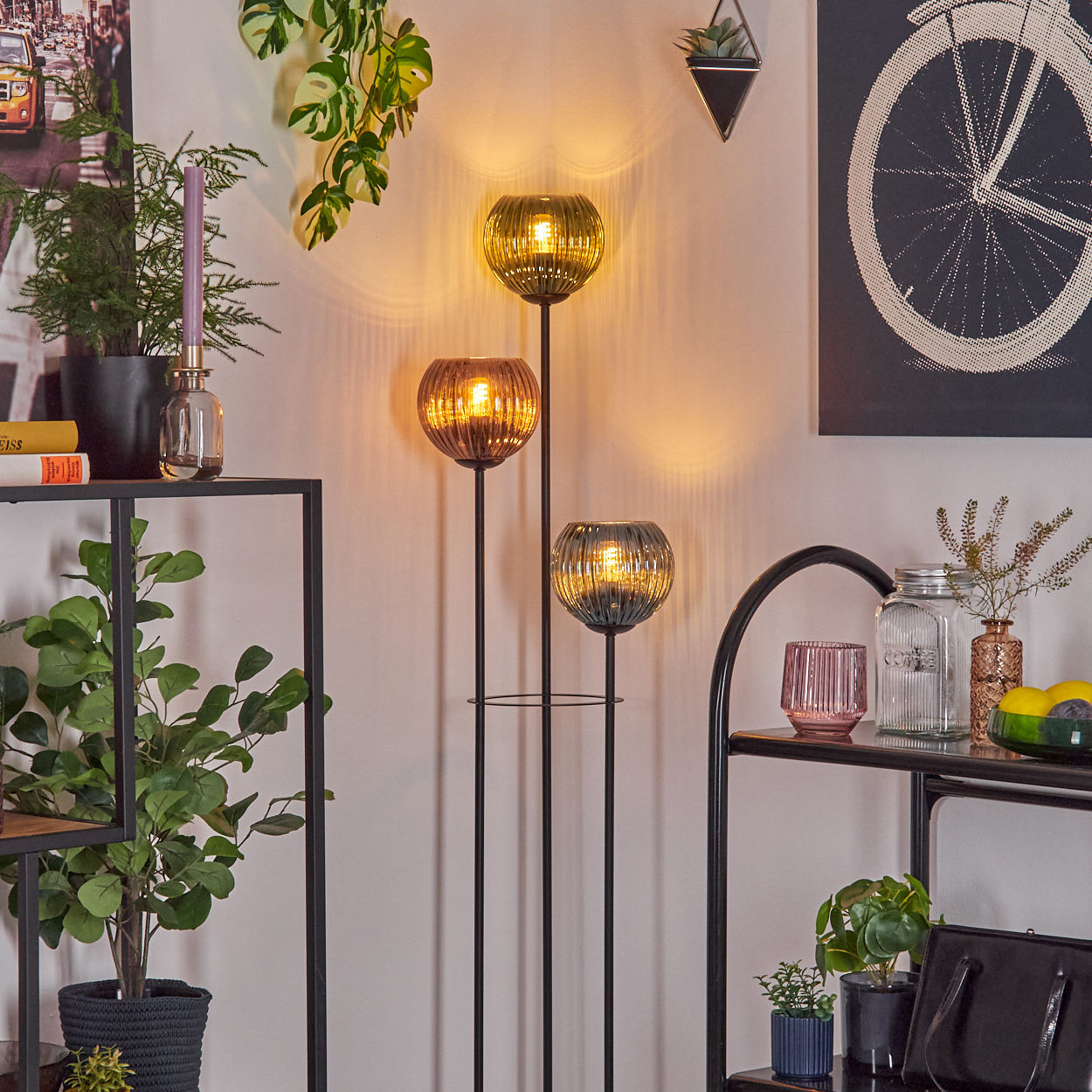 Stehlampe mit drei Lichtern und Glasschirmen, Pflanzen, Regalen und einem Fahrradrad-Kunstwerk.