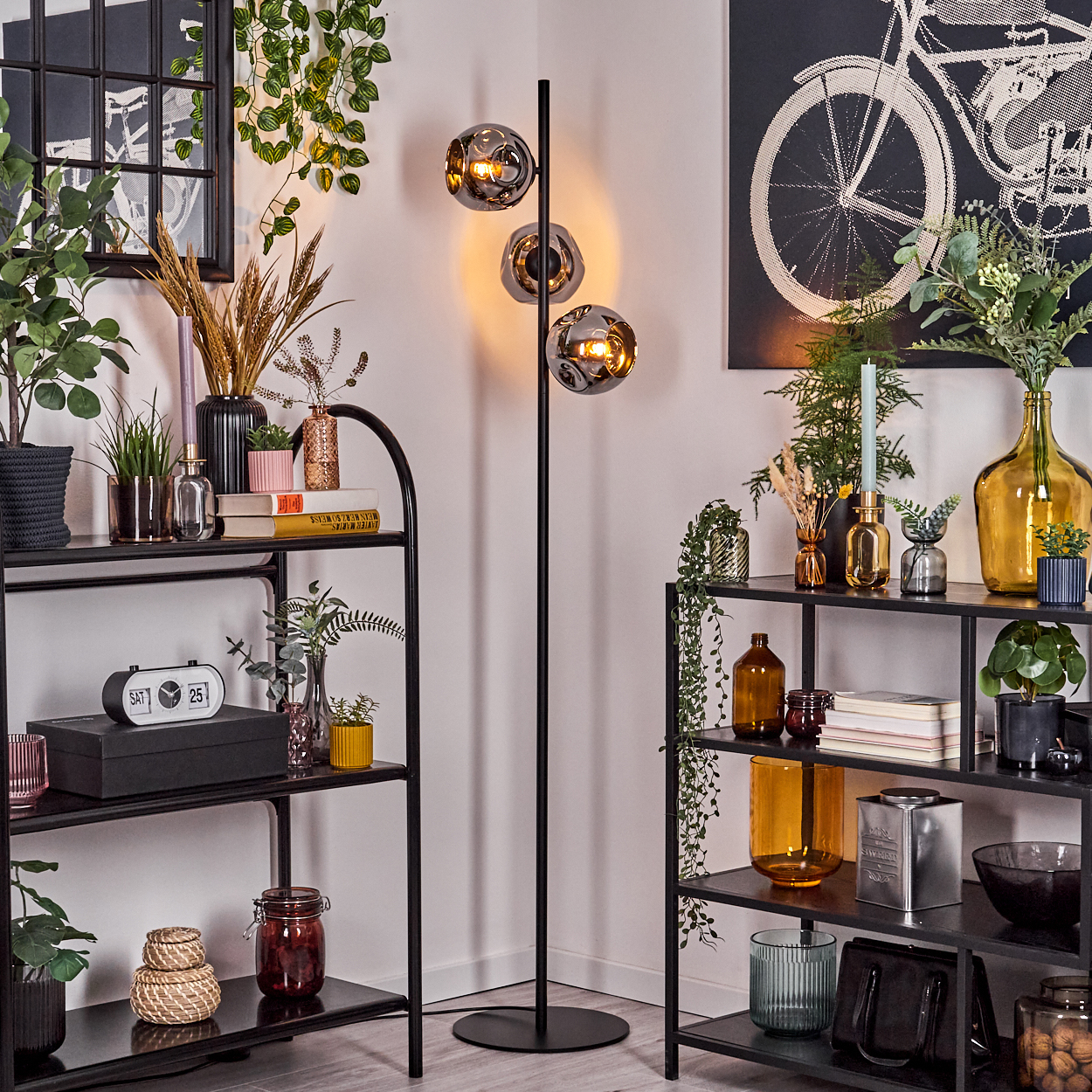 Ein Raum mit Regalen, einer Stehlampe und Wandkunst. Regale zeigen Pflanzen und Dekor. Die Lampe hat drei Glaskugeln.