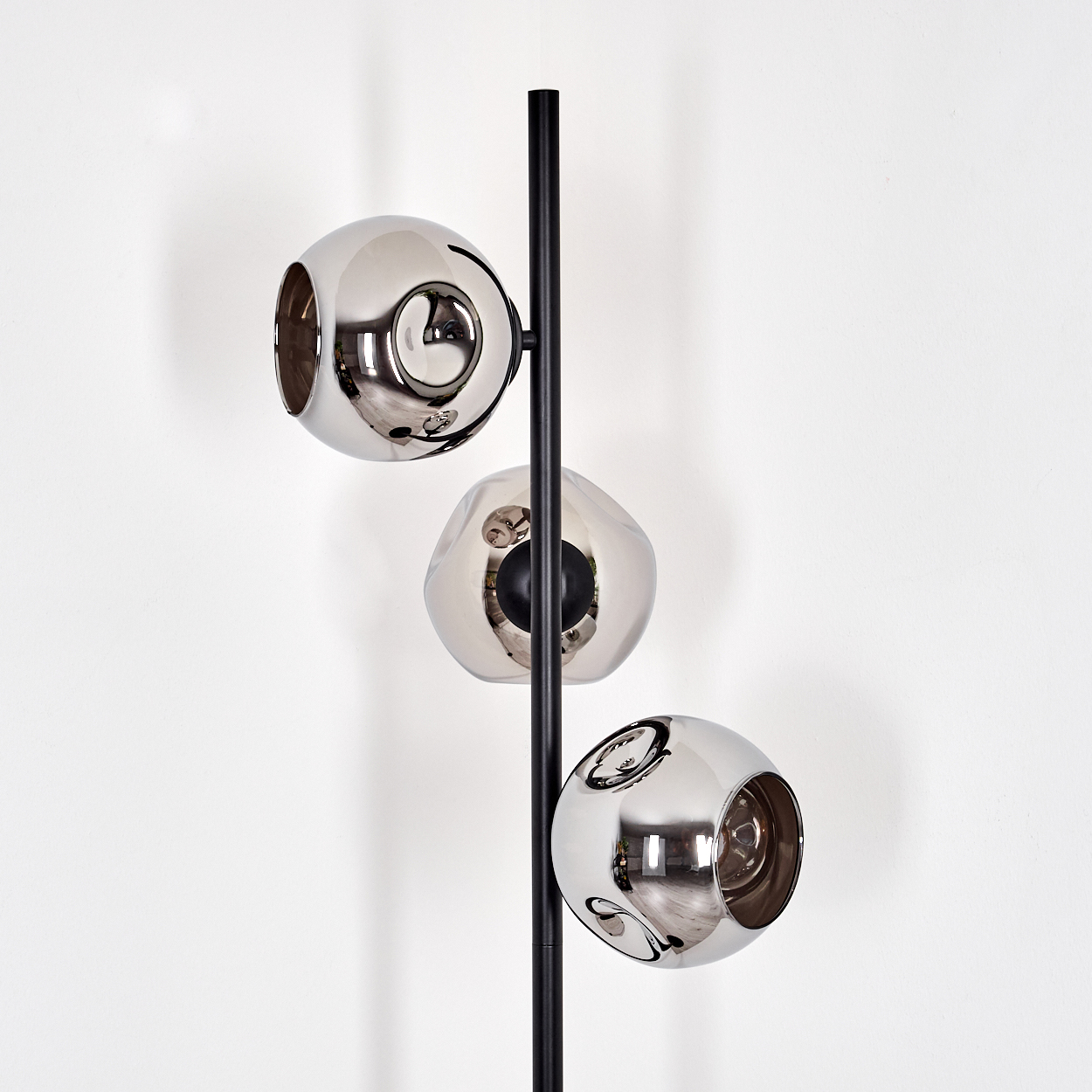 Eine moderne Lampe mit drei kugelförmigen, verspiegelten Schirmen. Ein schwarzer Pfosten trägt die Schirme vor einer weißen Wand.