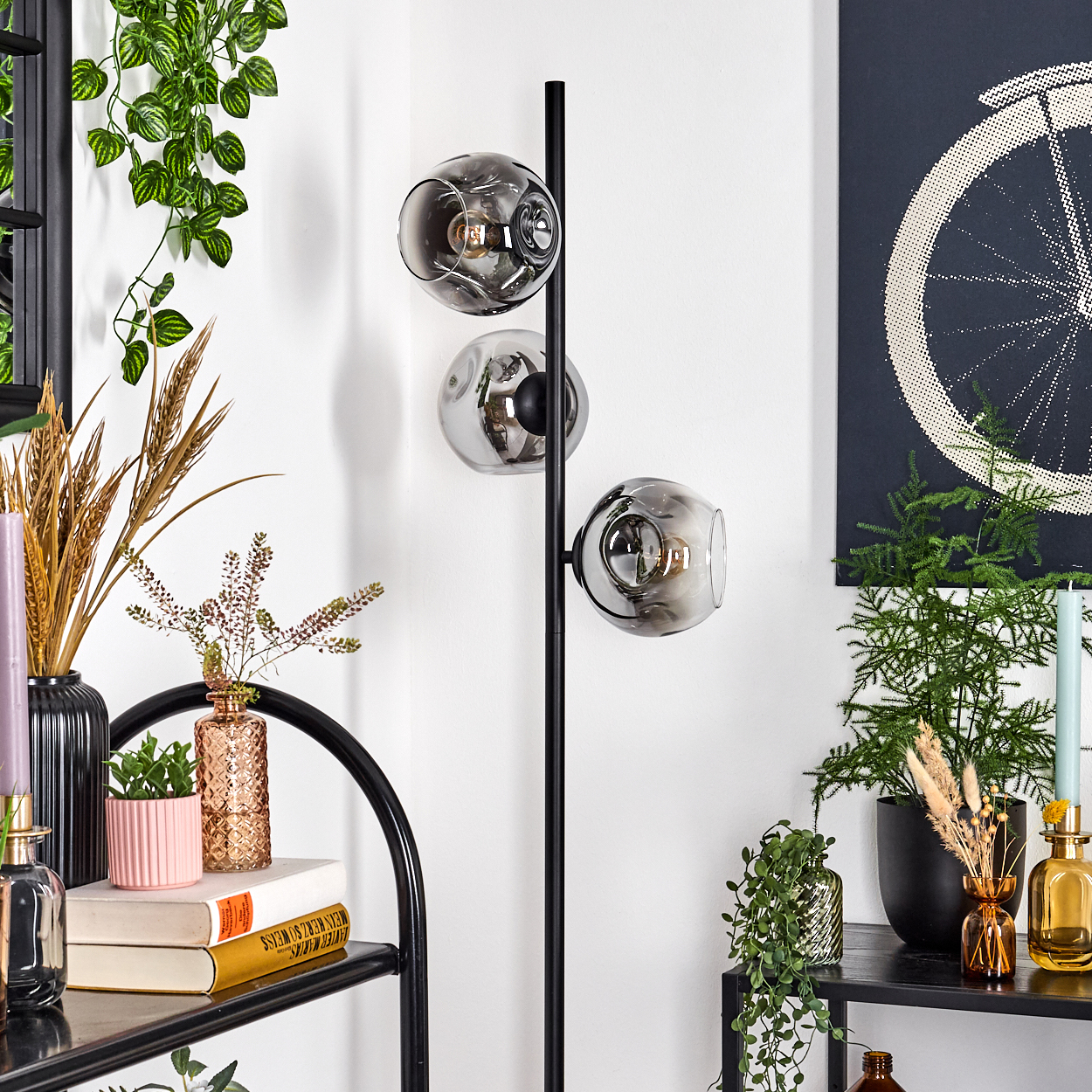 Ein modernes Zimmer mit einer schwarzen Stehlampe mit drei kugelförmigen Lichtern. Verschiedene Pflanzen, Vasen und Bücher sind auf Regalen angeordnet.