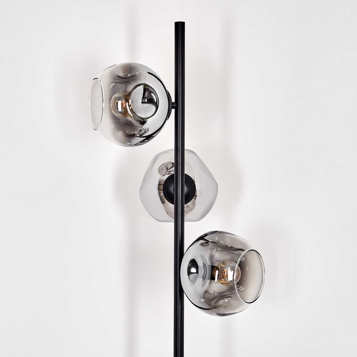 Eine moderne Lampe mit drei grauen Glaskugeln, jede mit einer Glühbirne. Die Lampe ist schwarz, vor einer weißen Wand.