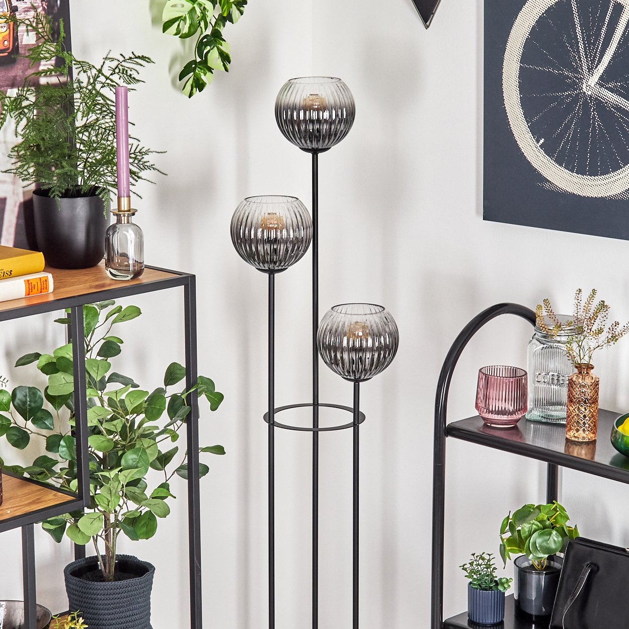 Eine dekorative Innenszene mit Glaskugel-Kerzenhaltern, Pflanzen und Regalen vor einer weißen Wand mit Kunstwerken.