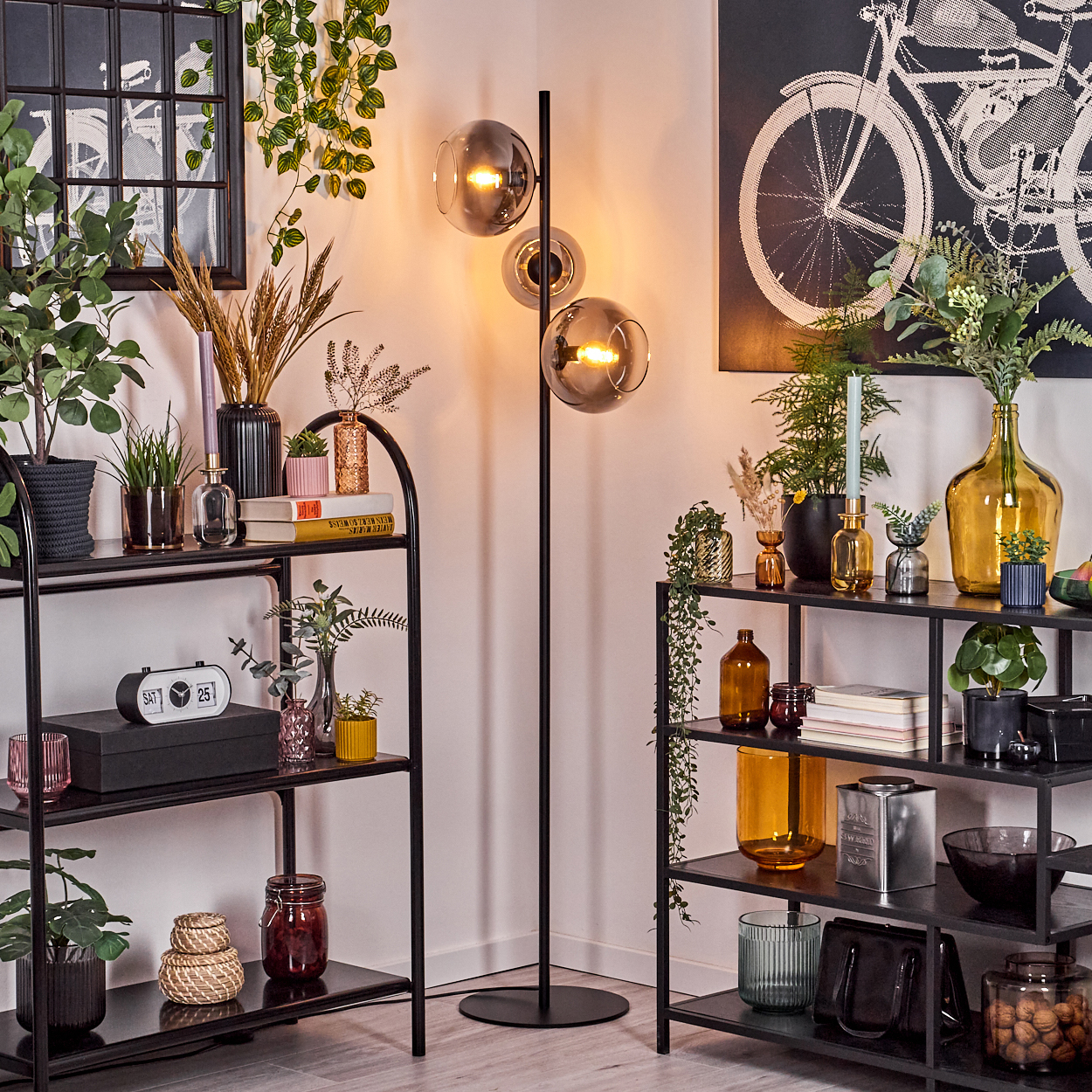 Eine Zimmerecke mit schwarzen Regalen, Pflanzen, einer Lampe und einem Fahrradkunstwerk an der Wand. Verschiedene Dekorationsartikel sind vorhanden.