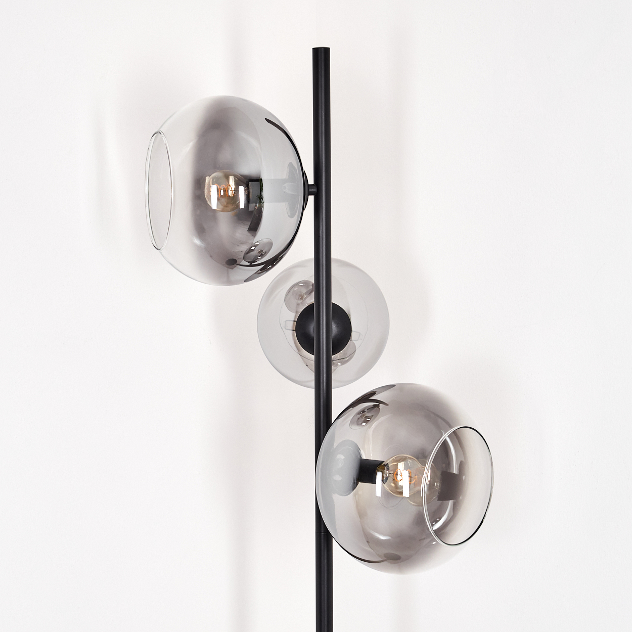 Eine moderne Stehlampe mit drei grauen Glaskugeln. Die Lampe hat einen schwarzen Metallrahmen. Der Hintergrund ist eine weiße Wand.