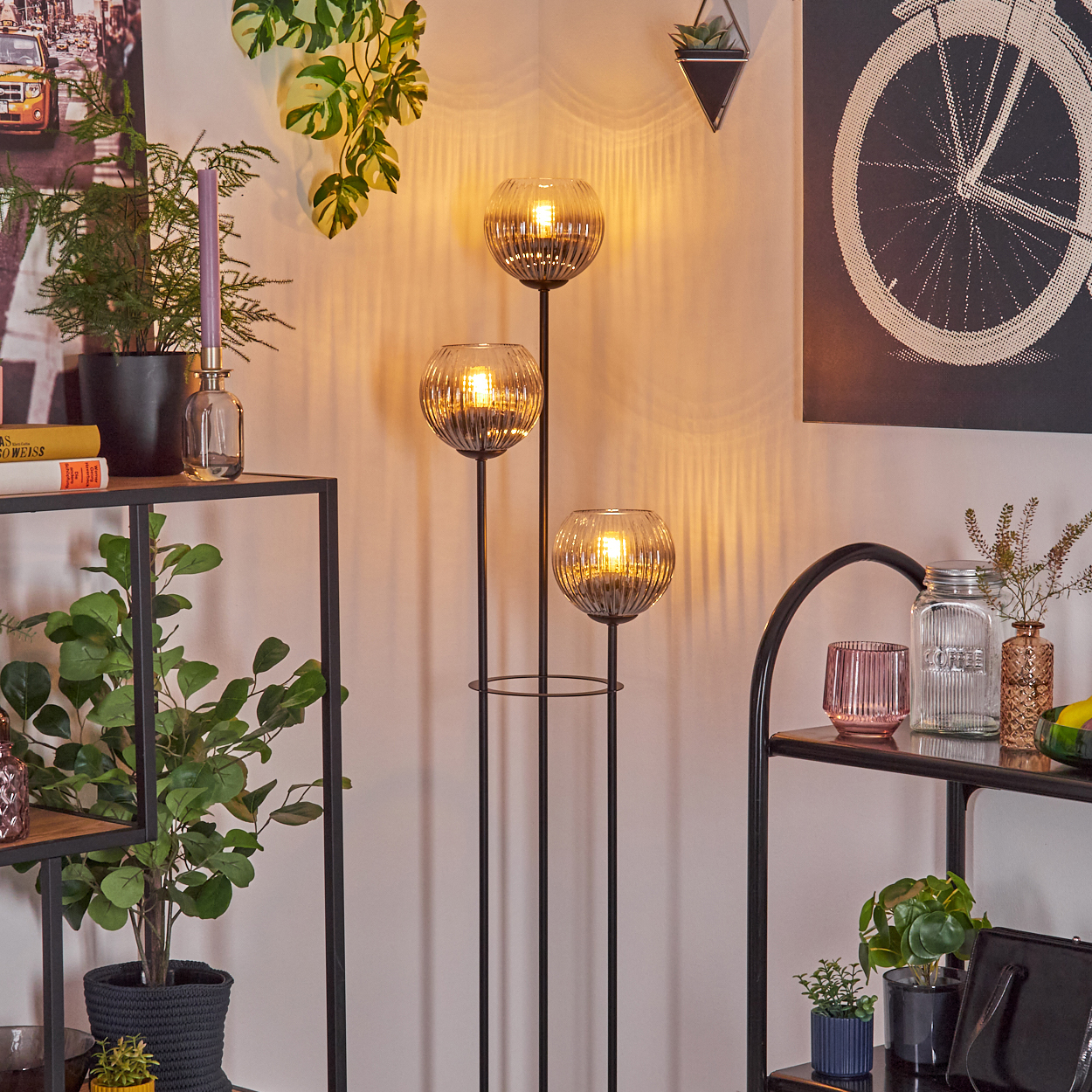 Eine Zimmerecke mit einer Stehlampe mit drei Glaskugeln, Pflanzen und einem schwarzen Metallregal mit Dekorationen.