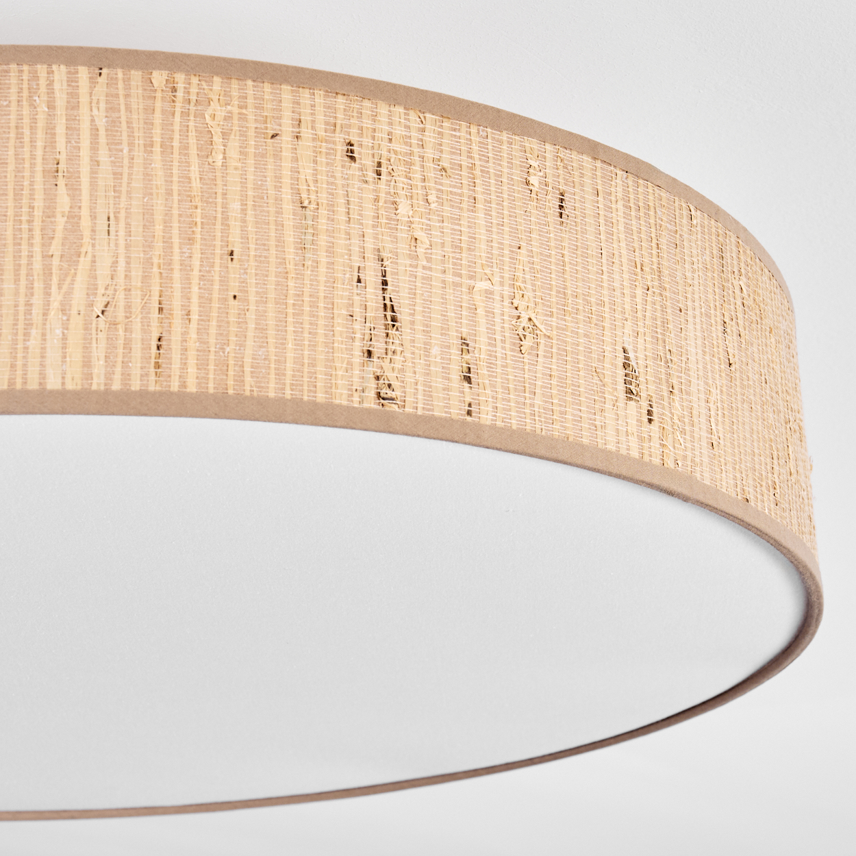 Nahaufnahme einer Deckenlampe. Sie hat einen gewebten, hellbraunen Schirm mit braunem Rand. Der Boden ist weiß.