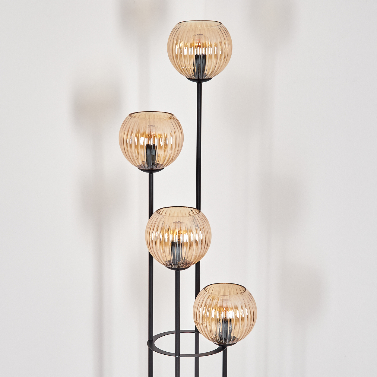 Eine moderne Stehlampe mit vier bernsteinfarbenen Glaskugeln. Die Lampe ist schwarz und steht vor einer weißen Wand.