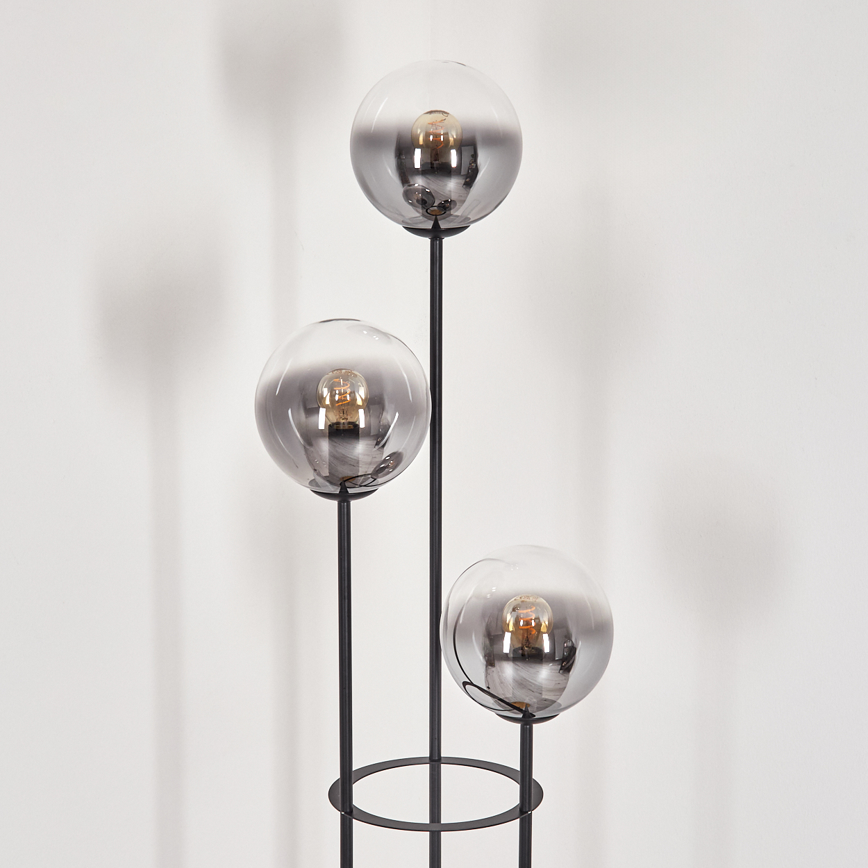 Moderne Stehlampe mit drei kugelförmigen Lampenschirmen. Jede Lampe hat eine Glühbirne. Die Lampe ist schwarz und steht vor einer weißen Wand.