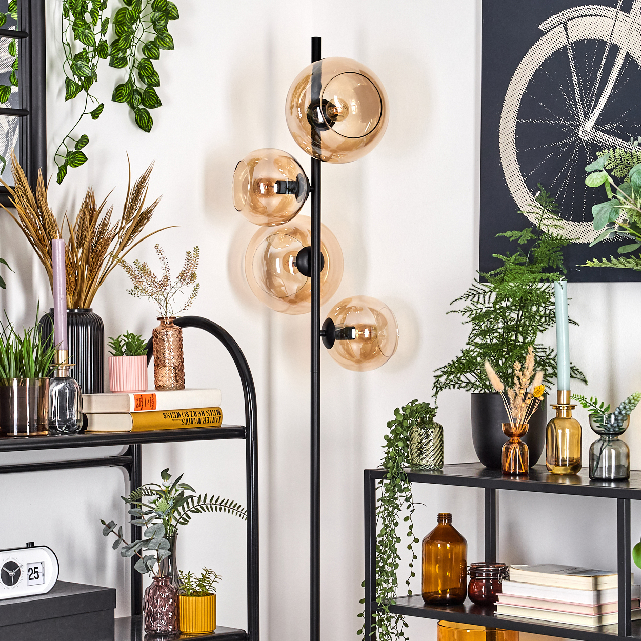 Eine Zimmerecke mit einer schwarzen Stehlampe mit Glaskugeln. Regale beherbergen Pflanzen, Bücher, Vasen und eine Uhr. Ein Fahrradrad-Kunstwerk ist an der Wand.