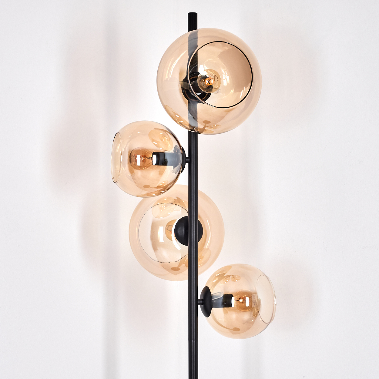 Eine moderne Stehlampe mit vier bernsteinfarbenen Glaskugeln auf einem schwarzen Metallrahmen. Der Hintergrund ist eine weiße Wand.