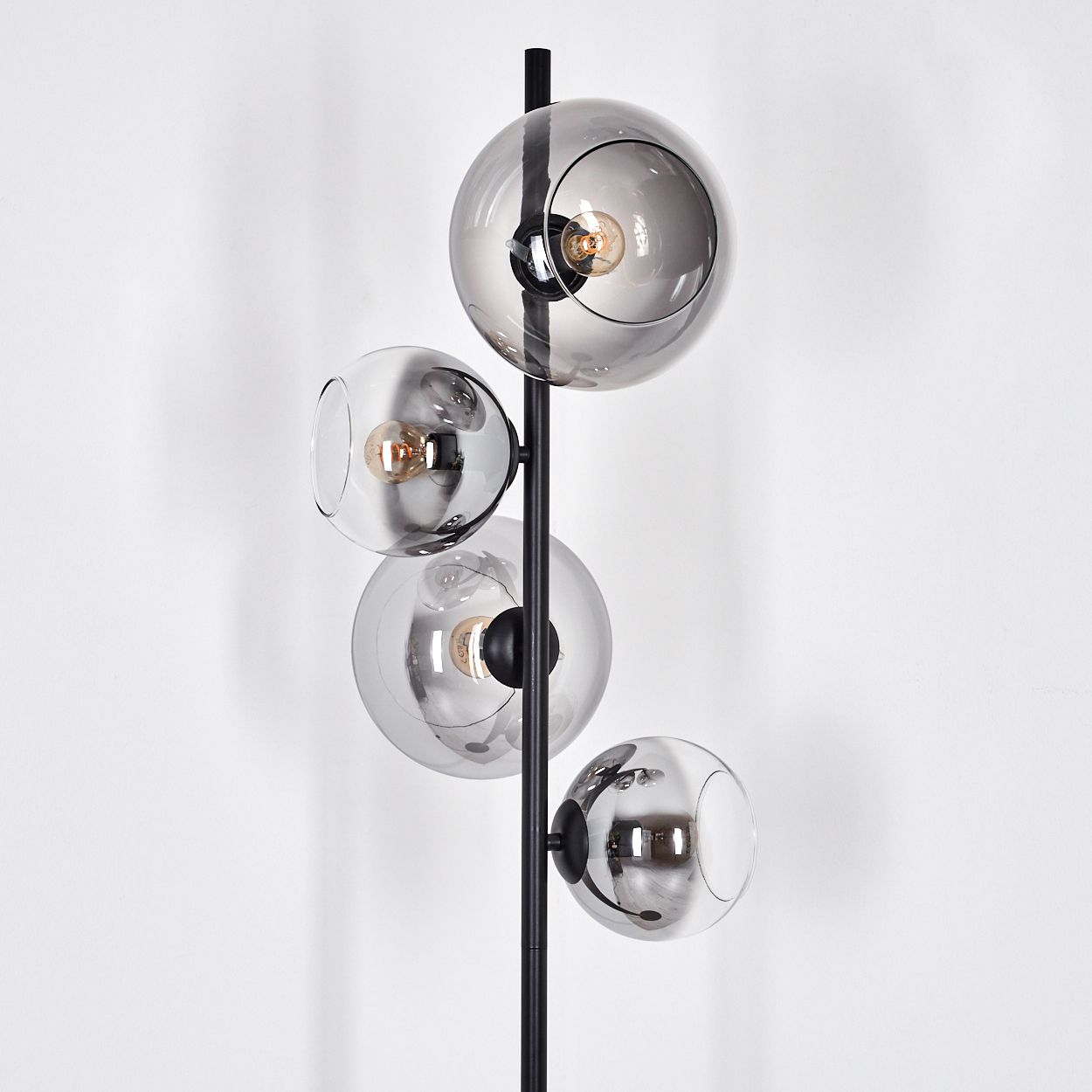 Eine moderne Stehlampe mit vier rauchgrauen Glaskugeln, jede mit einer Glühbirne, an einer schwarzen Metallsäule vor einer weißen Wand.