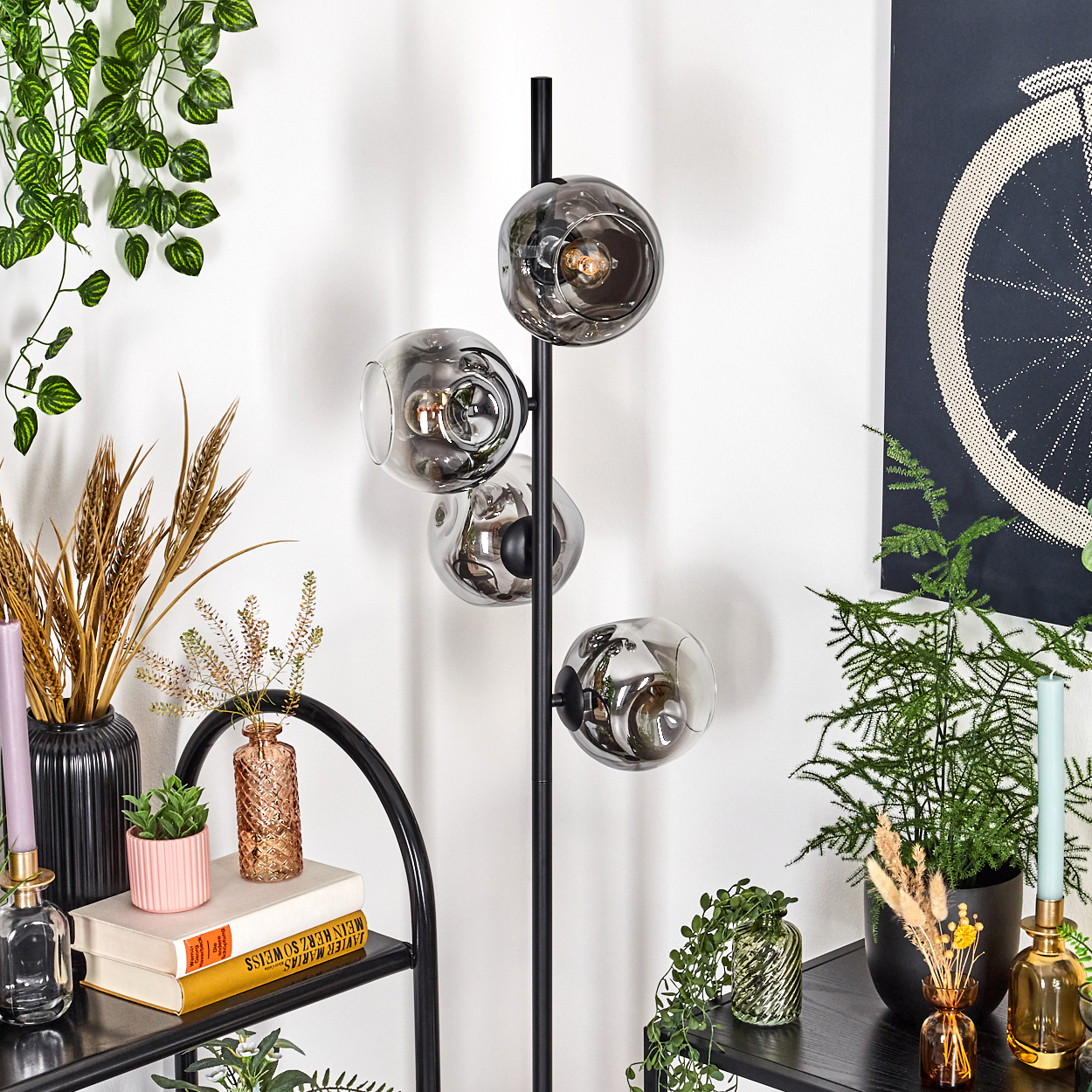 Eine moderne Stehlampe mit vier rauchfarbenen Glaskugeln, umgeben von Pflanzen, Kerzen und Dekorationsartikeln auf Regalen. Ein Fahrradrad-Kunstwerk.