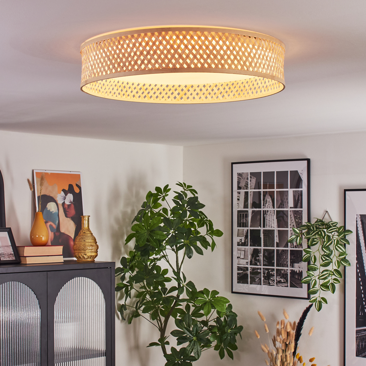 Ein Raum mit einer gewebten Deckenlampe. Ein Schrank mit Dekoration und eine große Pflanze. Kunstwerke und ein schwarz-wei ß Stadtbild Foto.