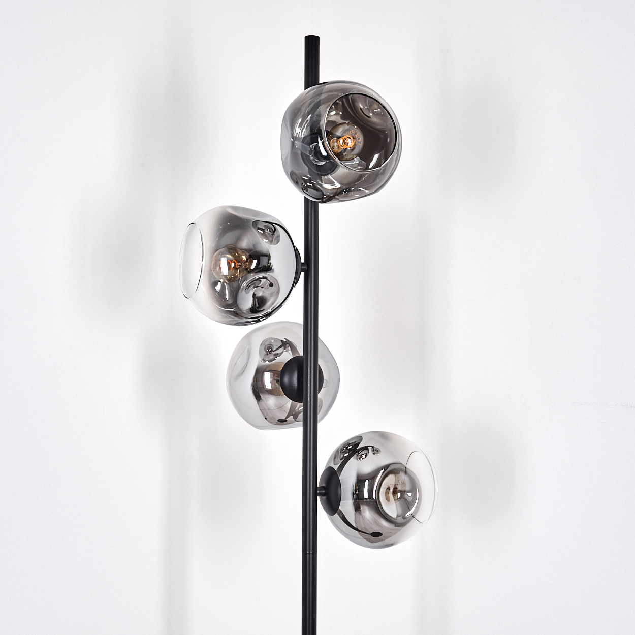 Eine moderne Stehlampe mit vier rauchgrauen Glaskugeln. Die Lampe ist schwarz und steht vor einer schlichten weißen Wand.