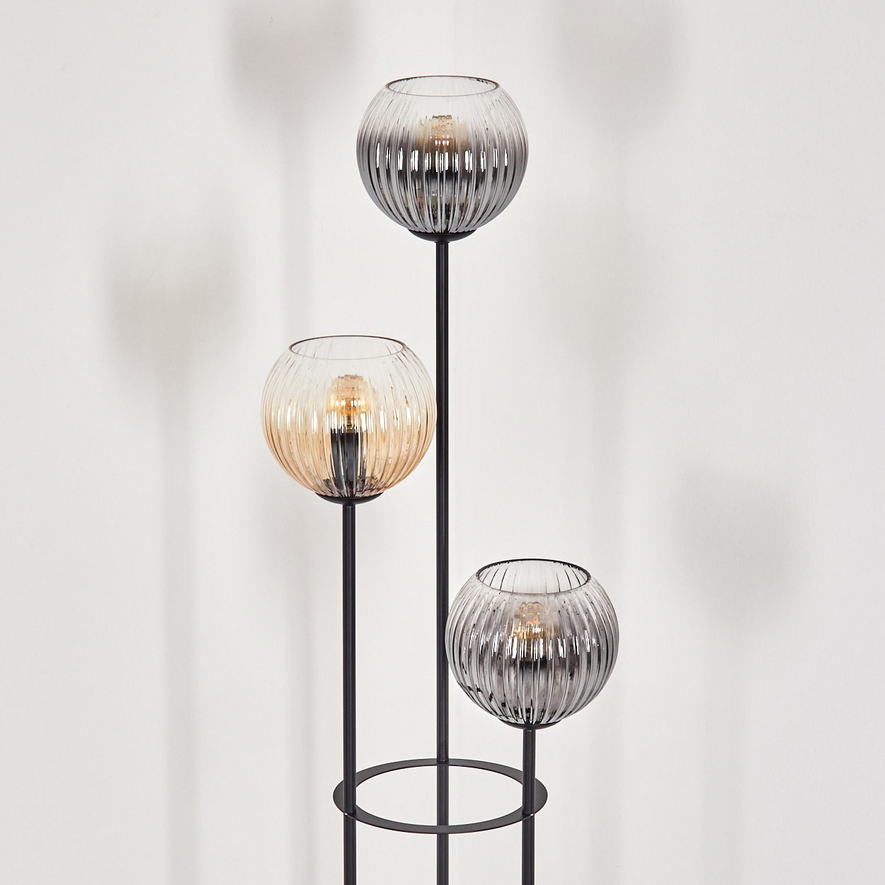 Eine moderne Lampe mit drei Glaskugeln. Jede Kugel hat eine Glühbirne. Basis und Stiele sind schwarz, vor einer weißen Wand.