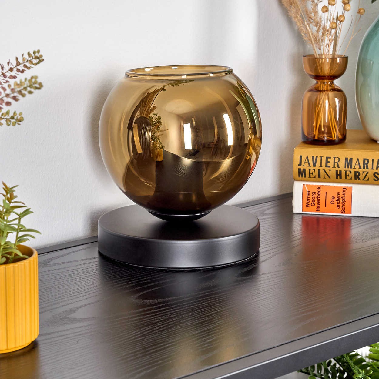 Eine goldene Glasschale steht auf einem schwarzen Sockel. Andere Vasen und Bücher stehen auf einer dunklen Holzoberfläche, mit einer kleinen Pflanze.