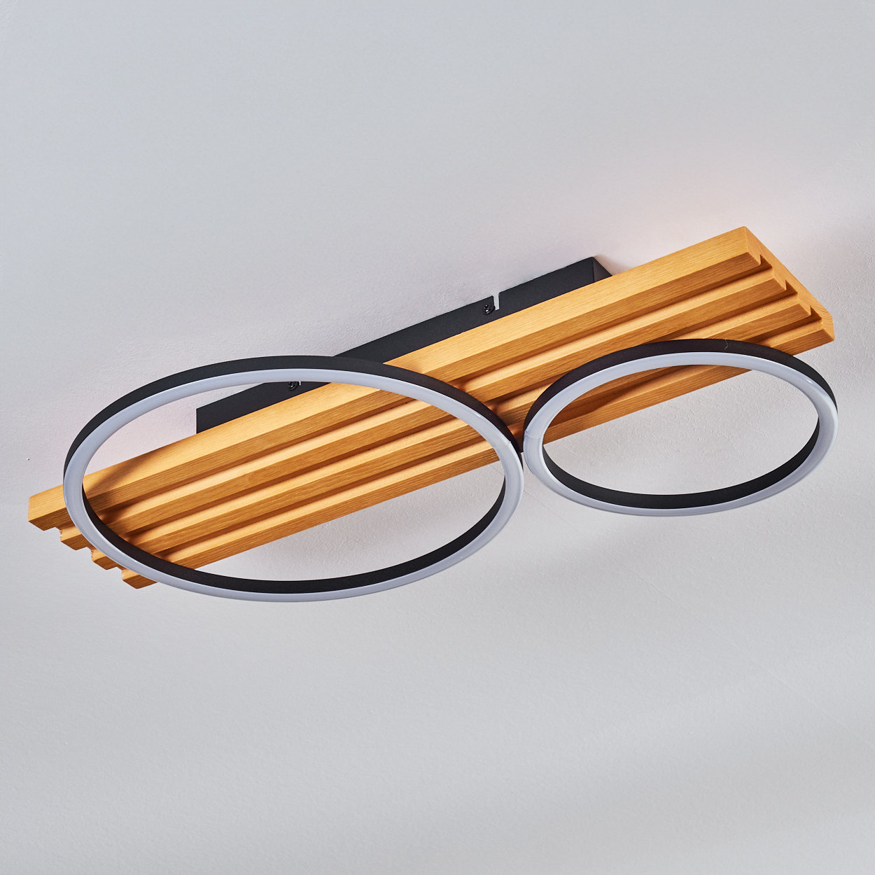 Deckenleuchte mit zwei Ringlichtern und Holzpaneel. Schwarz-weiße Ringe, Holzmaserung, an einer weißen Decke montiert.