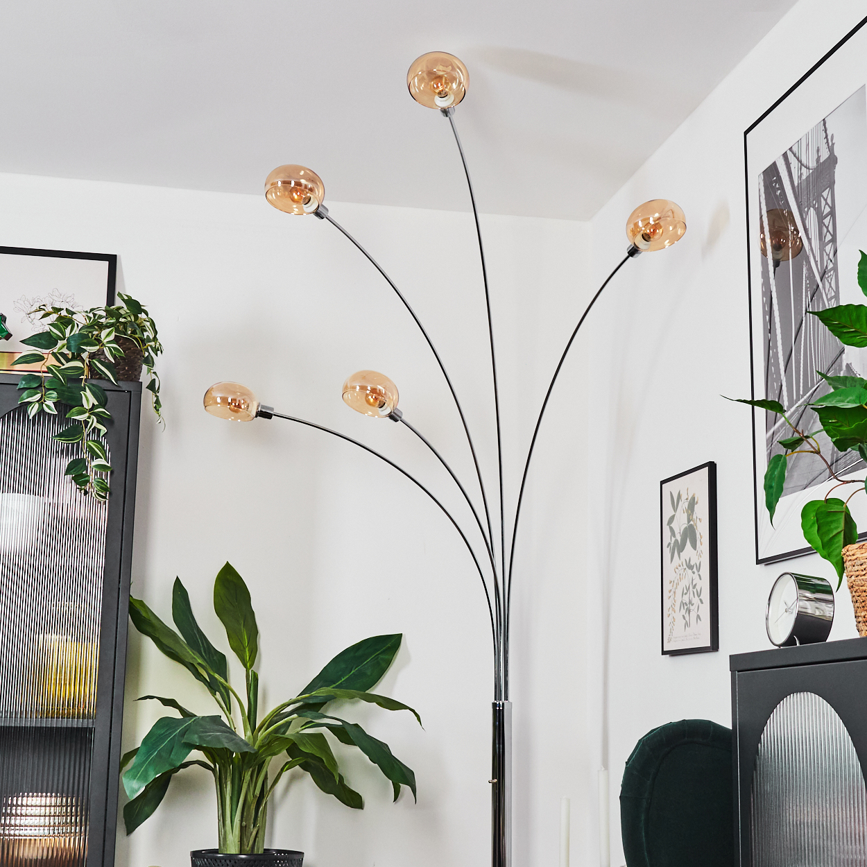 Eine moderne Stehlampe mit fünf Glühbirnen. Der Raum hat Pflanzen, schwarze Möbel und gerahmte Bilder. Die Wände sind weiß.