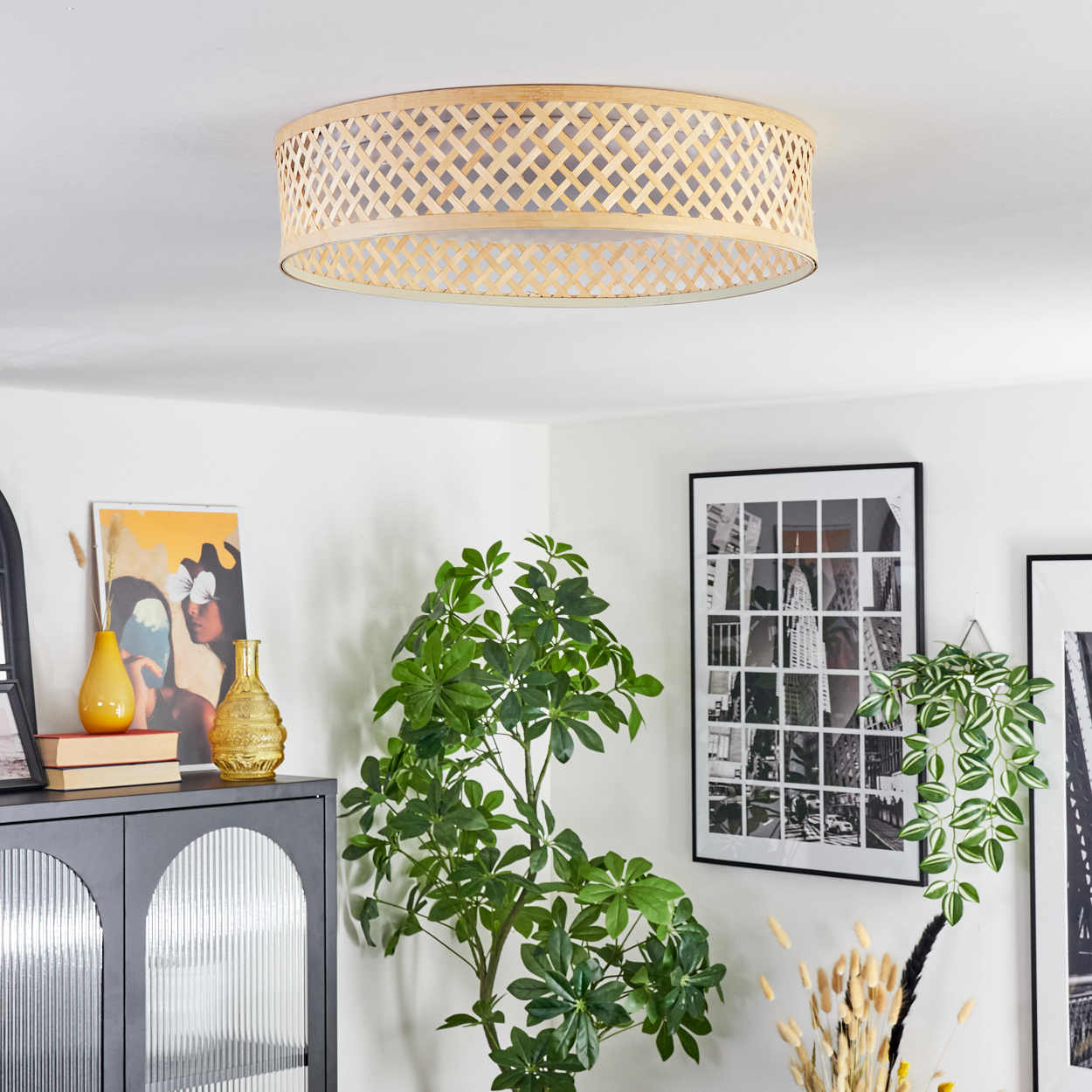 Ein Raum mit einer gewebten Deckenleuchte, einem schwarzen Schrank, einer Pflanze, gerahmten Bildern und einer gelben und goldenen Vase.