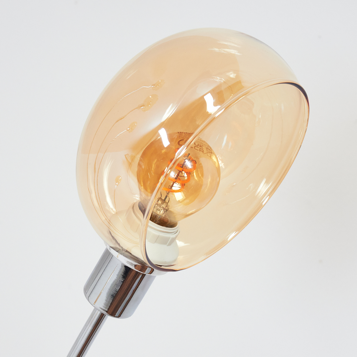 Nahaufnahme einer Lampe. Sie hat einen Chromschaft, eine Glühbirne und einen Glasschirm. Die Glühbirne leuchtet orange, der Schirm ist bernsteinfarben.
