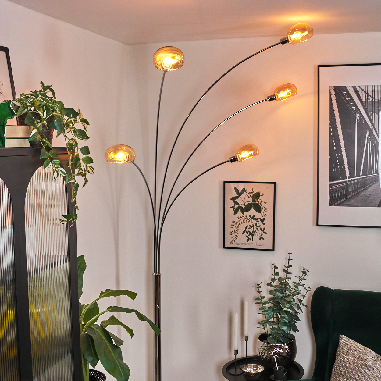 Ein modernes Wohnzimmer mit Stehlampe, Pflanzen, Kunstwerken und einem grünen Sessel. Die Lampe hat fünf bernsteinfarbene Glasschirme.