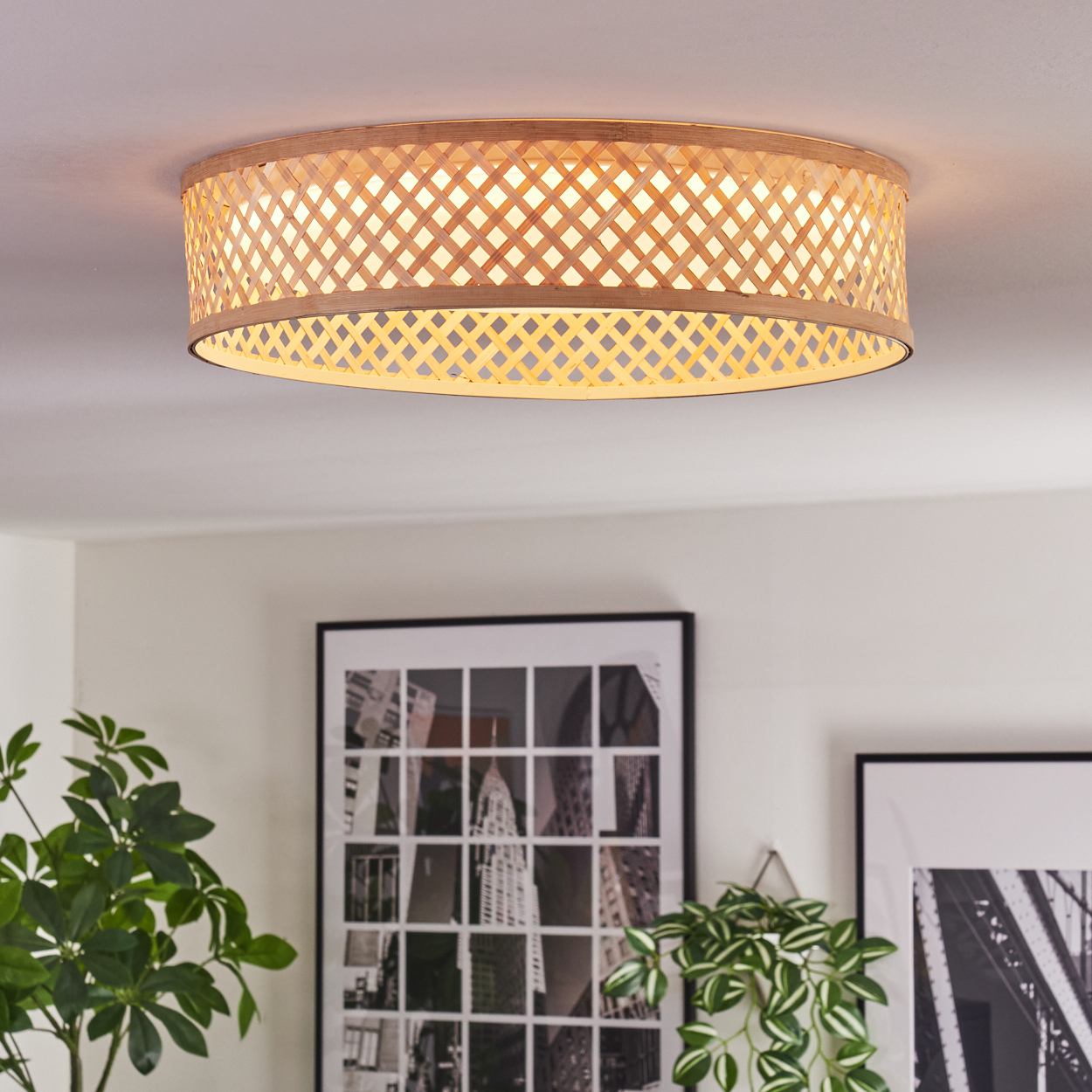 Eine gewebte Deckenlampe aus Bambus hängt an der Decke. Darunter befinden sich gerahmte Bilder und Pflanzen. Die Lampe ist beleuchtet.