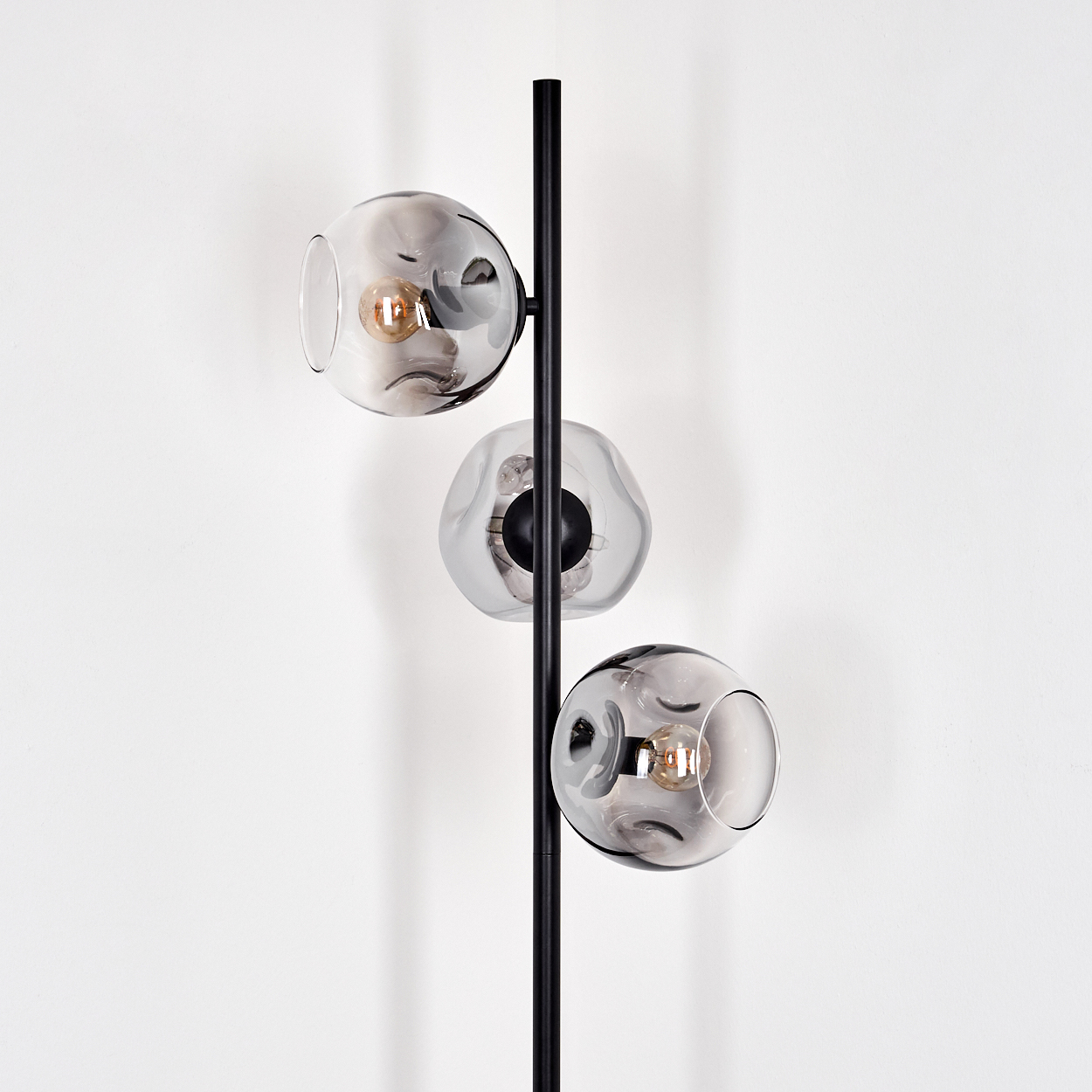Eine moderne Stehlampe mit drei kugelförmigen, rauchfarbenen Glasschirmen. Die Lampe hat eine schwarze Metallsäule vor einer weißen Wand.