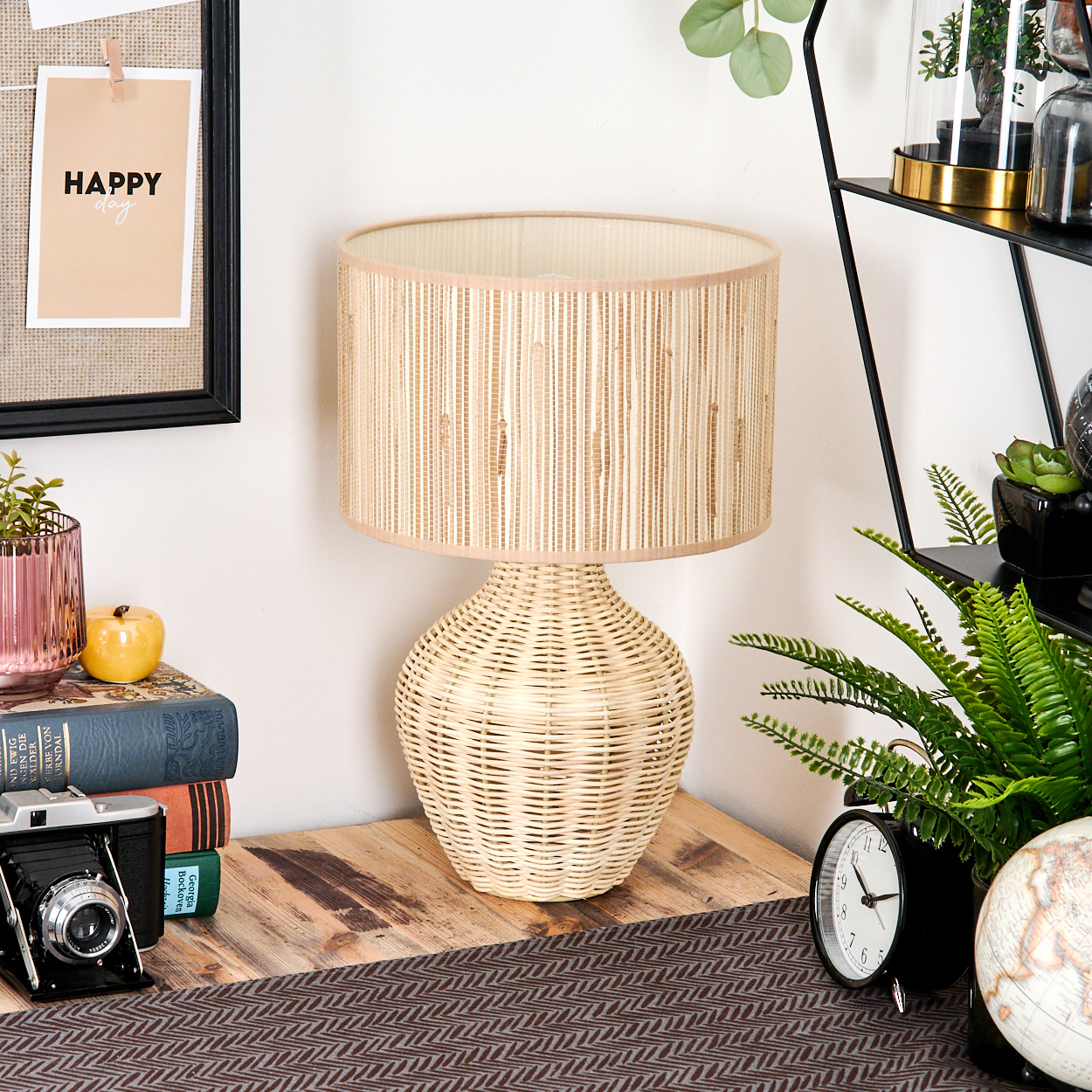 Eine gewebte Lampe steht auf einem Holztisch mit Büchern, einer Kamera und einer Uhr. Eine Pinnwand mit einem 'Happy Day'-Zettel ist sichtbar.