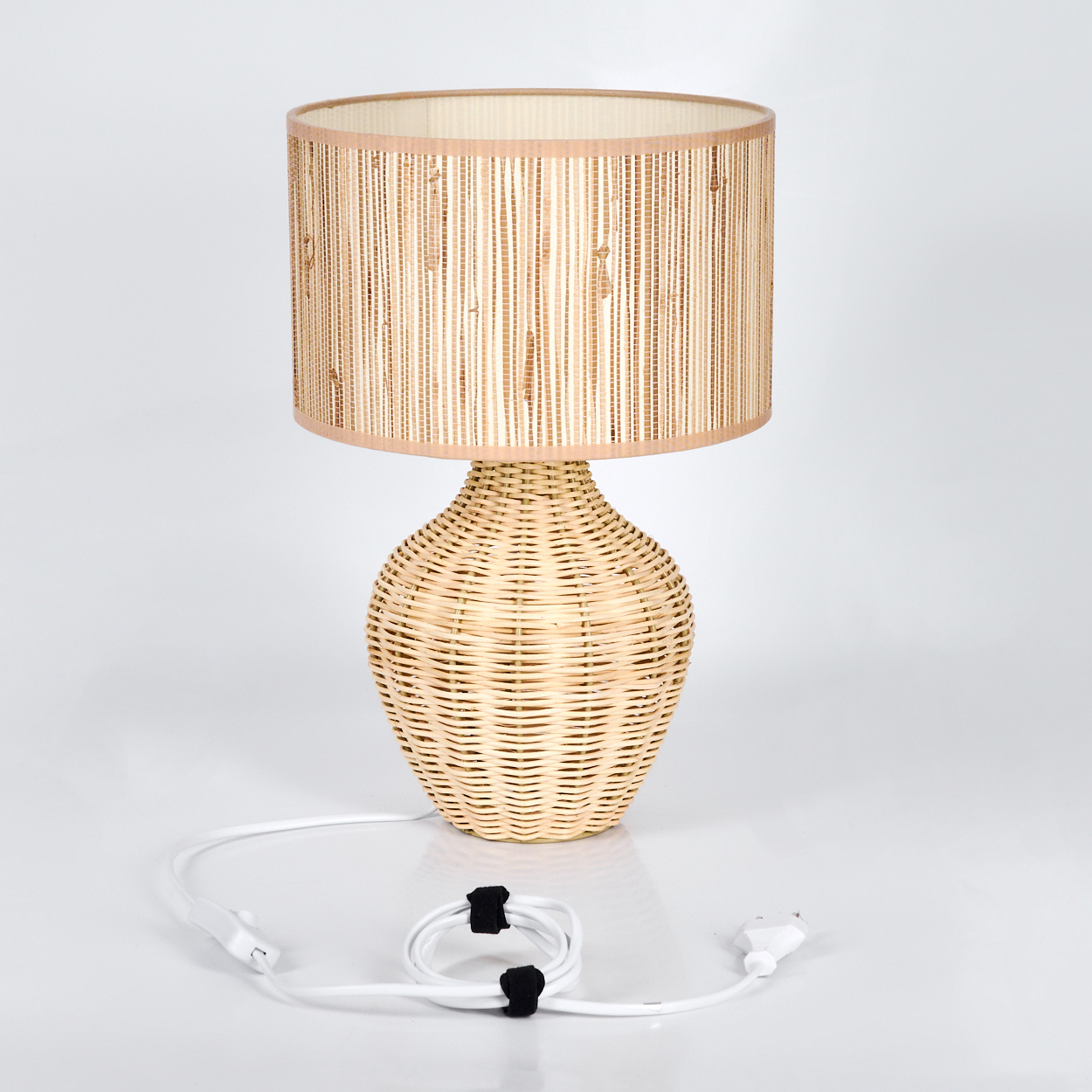 Eine gewebte Korblampe mit beigem Lampenschirm. Ein weißes Kabel mit Stecker und schwarzen Kabelbindern befindet sich vor der Lampe.
