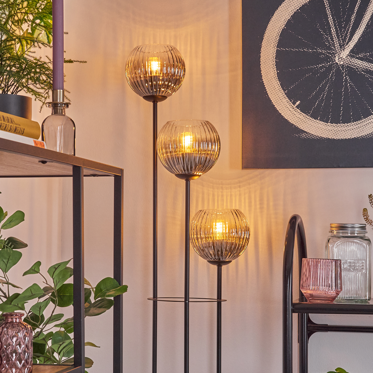 Eine dekorative Szene mit einer dreiflammigen Stehlampe, Regalen mit Dekoration und einem gerahmten Kunstdruck eines Fahrradrads.