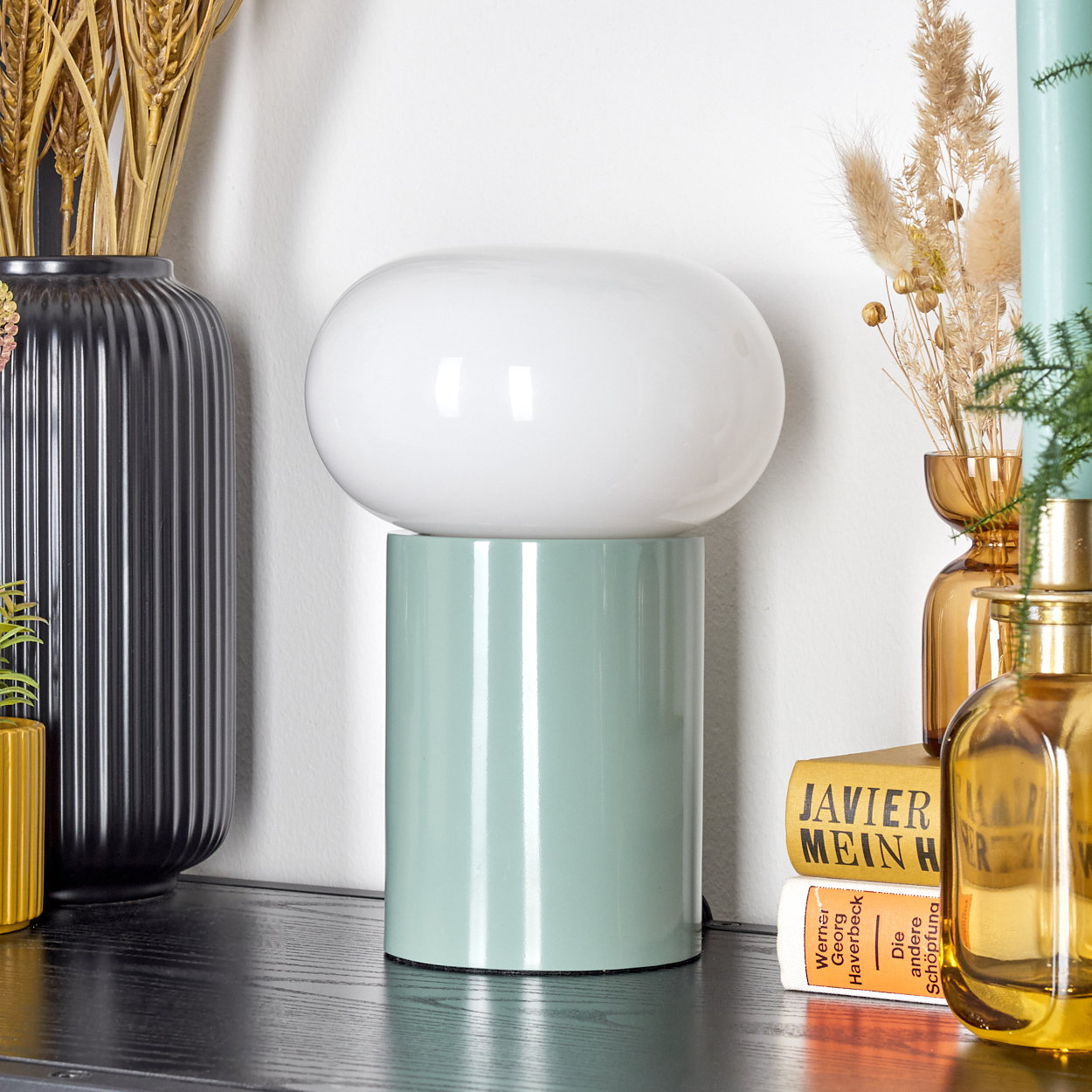 Eine weiße Lampe auf mintgrüner Basis steht auf einer schwarzen Oberfläche mit Vasen und Büchern. Hintergrund mit Weizen und goldenen Vasen.